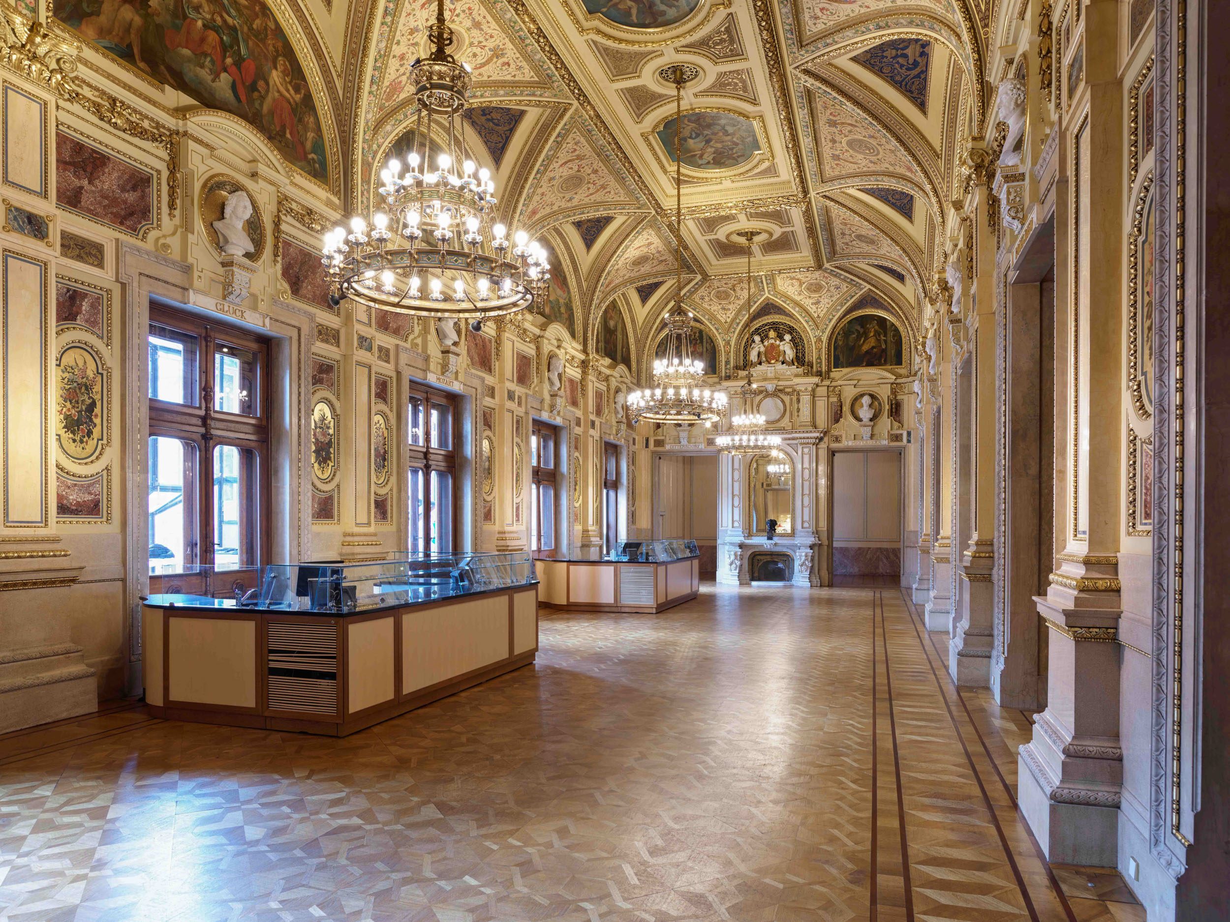 A grand, elegantly decorated room with high ceilings featuring intricate patterns and chandeliers. Large windows allow natural light to illuminate the space, which includes wooden floors and decorative moldings. Two reception desks with glass display cases are positioned side by side.