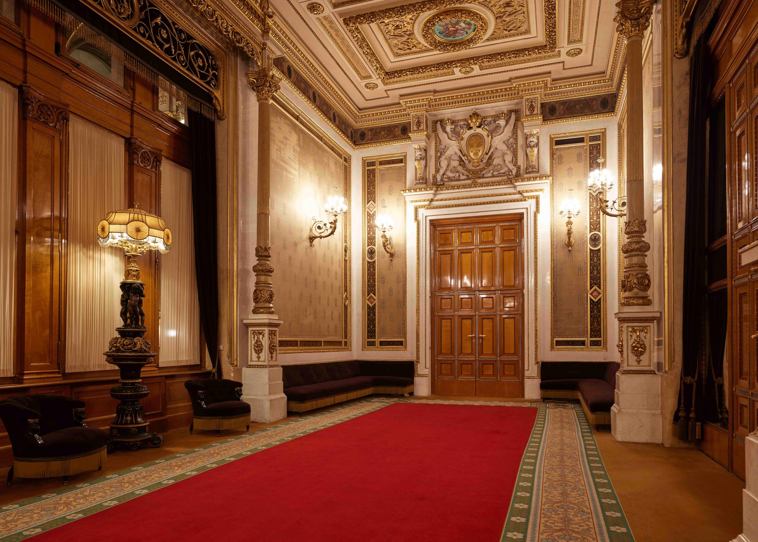 Der Raum ist opulent eingerichtet, mit einem roten Teppich und elegantem Holzmobiliar. An den Wänden befinden sich kunstvolle Verzierungen und Beleuchtung. Eine nostalgische Stehlampe und bequeme Sessel laden zum Verweilen ein. Eine große Holztür in der Mitte führt zu einem anderen Bereich des Raumes.