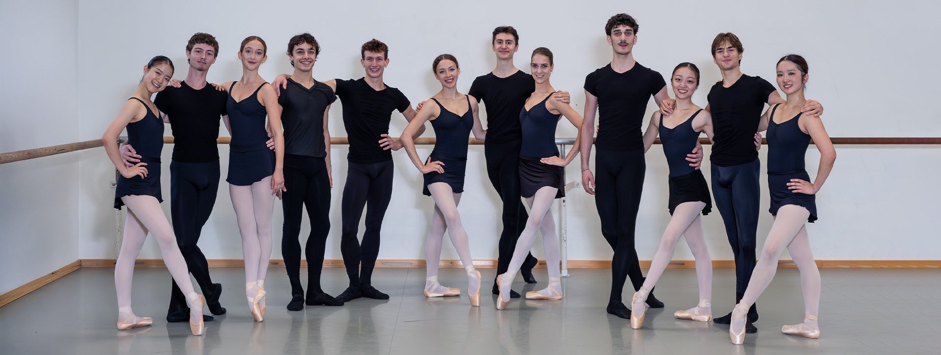 Eine Gruppe von zwölf Balletttänzern steht in einem Tanzstudio. Sie tragen schwarze Oberteile und verschiedene Skirts oder Leggings. Einige tragen Spitzenschuhe. Alle lächeln in die Kamera und posieren in einer freundlichen Haltung. Im Hintergrund ist eine Tanzstange sichtbar.
