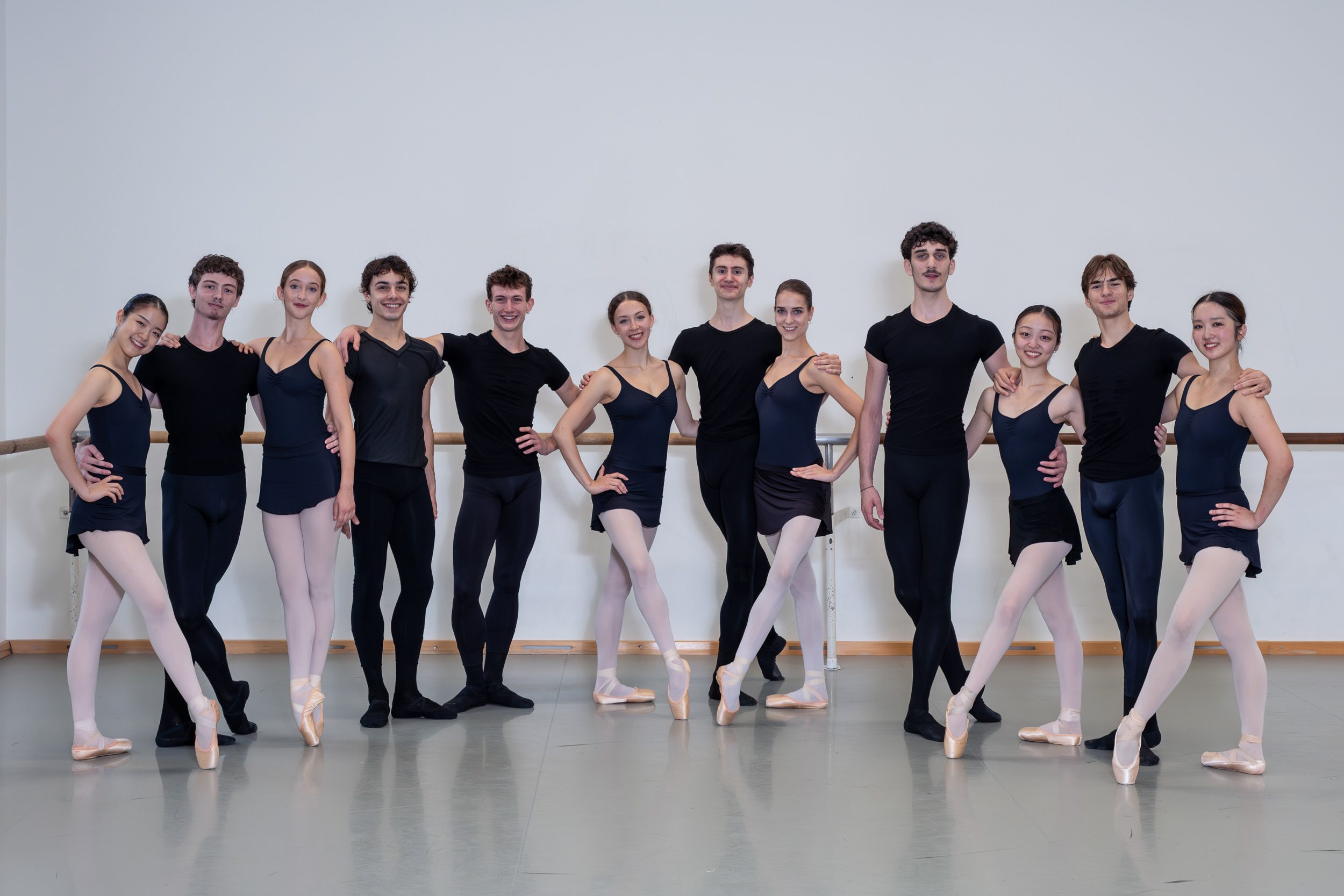Eine Gruppe von 12 Balletttänzern, männlich und weiblich, steht gemeinsam in einer Tanzschule in engem Kreis. Sie tragen schwarze Ballettkleidung und einige haben Spitzenschuhe an. Alle posieren lächelnd, mit einem harmonischen Hintergrund aus hellen Wänden und einem Bodenbelag zum Tanzen.