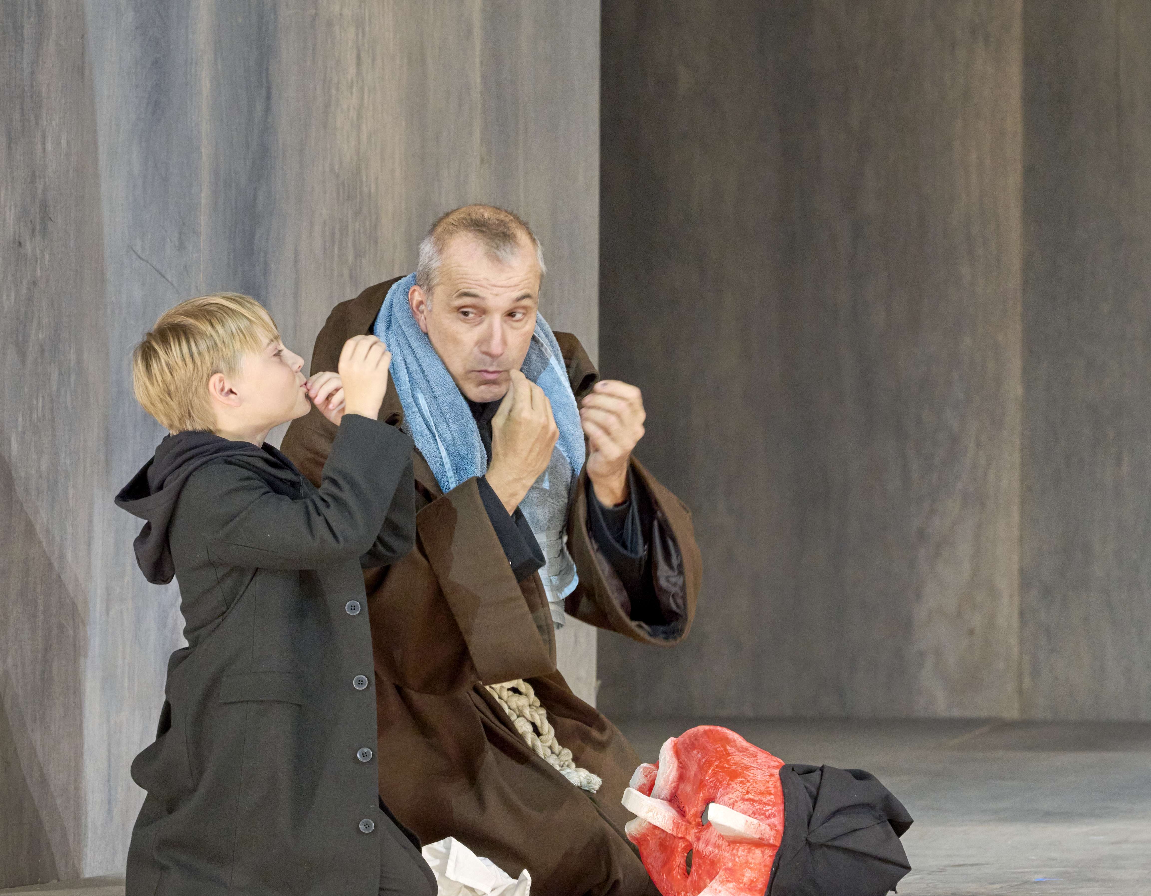 Ein Mann in einem braunen Gewand sitzt auf einer Bühne und schaut aufmerksam auf einen Jungen, der neben ihm steht. Der Junge hat kurze, blonde Haare und trägt einen schwarzen Mantel. Beide scheinen in ein Gespräch vertieft zu sein.