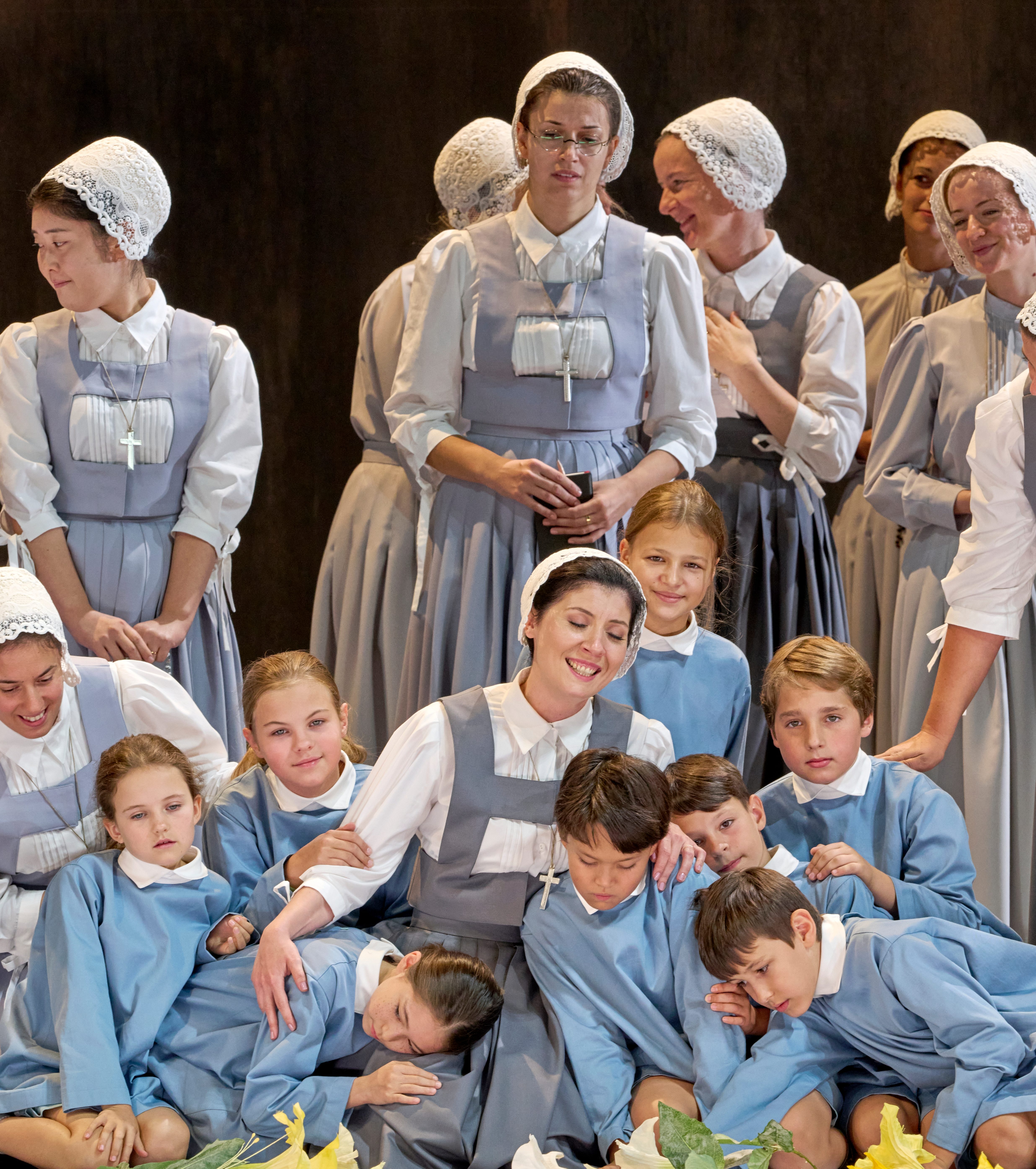 Eine Gruppe von Frauen und Kindern in traditionellen, grauen Kleidern steht zusammen. Eine Frau sitzt im Mittelpunkt und umarmt mehrere Kinder, die entspannt an ihr lehnen. Die Szene strahlt Gemeinschaft und Fürsorglichkeit aus.