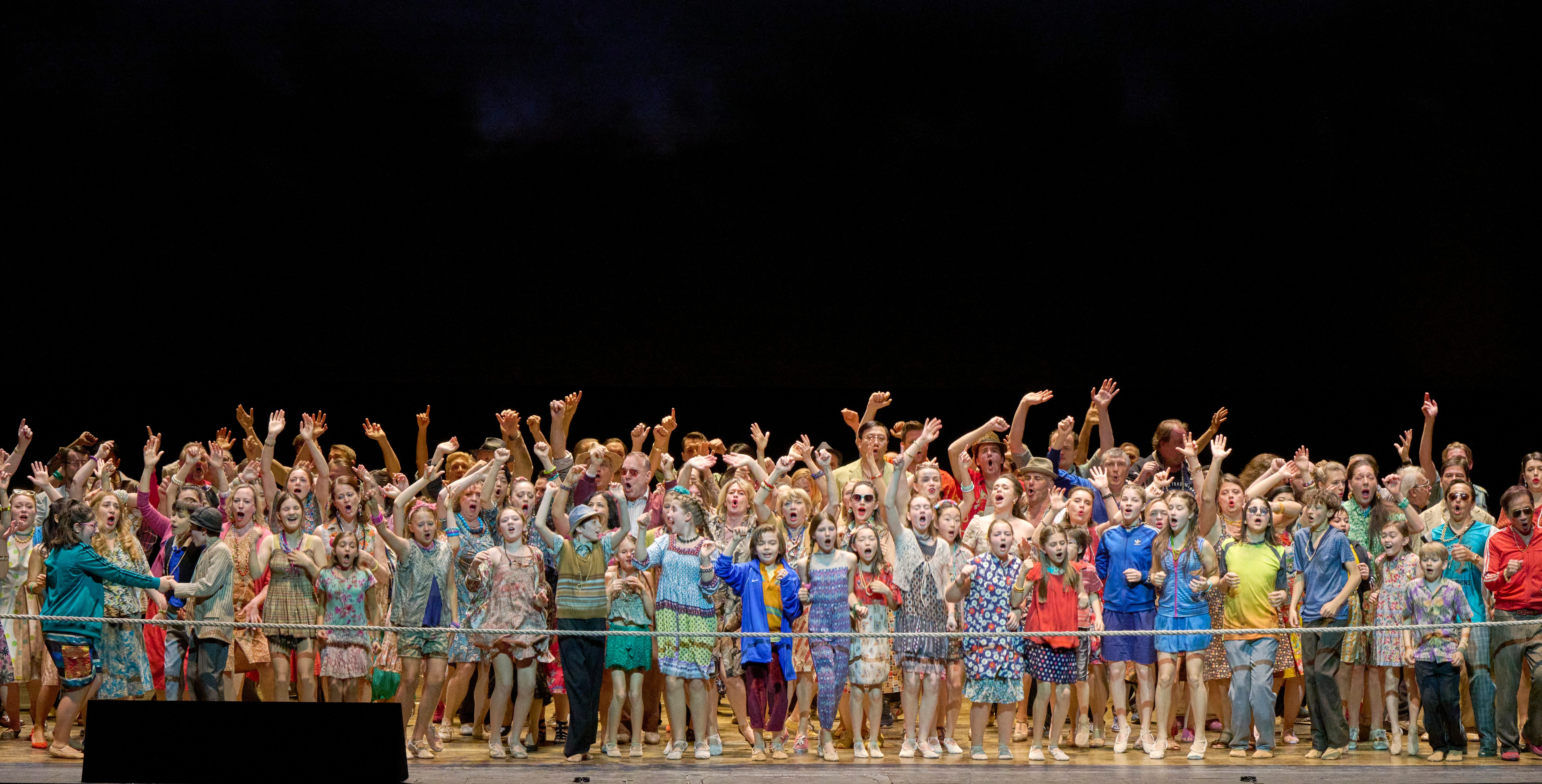 Eine große Gruppe von Menschen steht auf einer Bühne und singt begeistert. Viele tragen bunte Kleidung und haben ihre Hände in die Luft erhoben. Die Stimmung ist fröhlich und festlich, während die Darsteller zusammen eine lebendige Aufführung präsentieren.