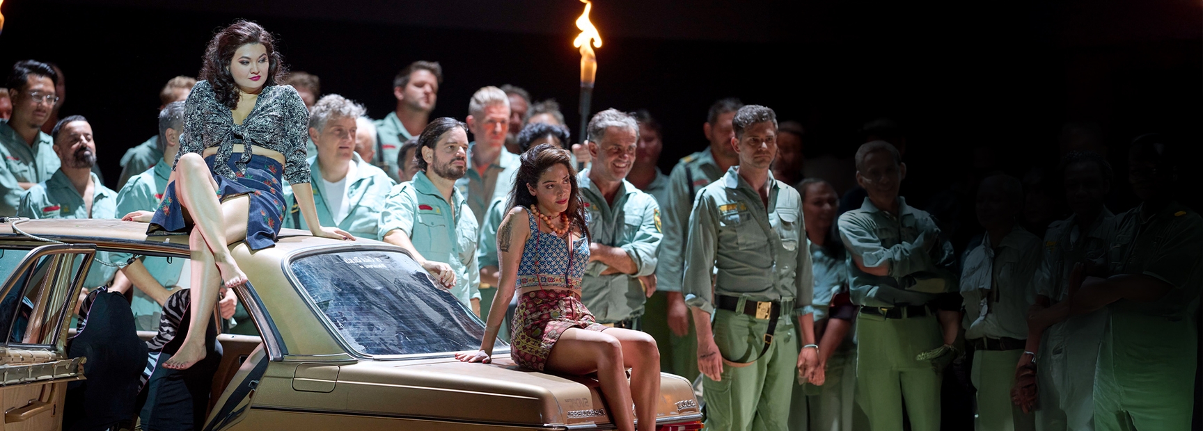 Two women in vibrant outfits sit on a vintage car, exuding confidence. Behind them, a group of men in uniforms watches intently, creating a backdrop of curiosity and tension. The scene suggests a performance filled with drama and intrigue.
