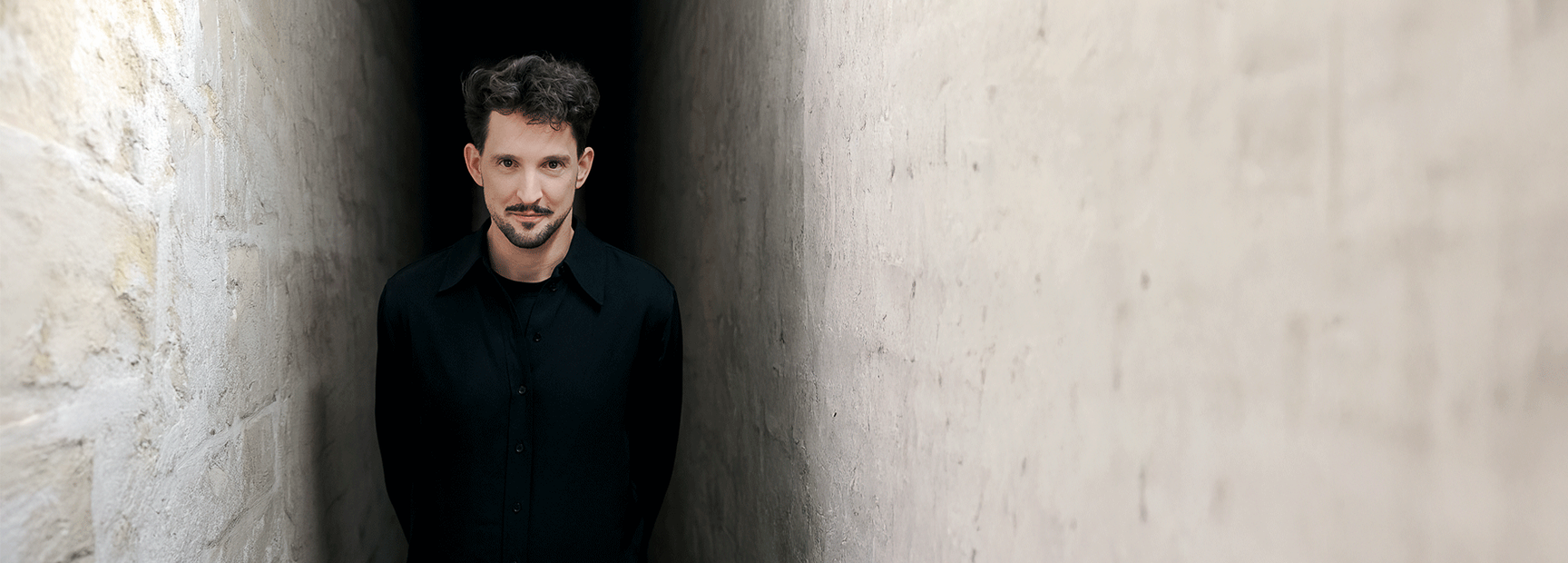 Ein Mann steht in einem engen, dunklen Durchgang zwischen zwei Wänden. Er trägt ein schwarzes Hemd, hat kurze, lockige Haare und einen leichten Bart. Sein Blick ist ernst, und er lehnt leicht nach vorne. Die Wände sind aus hellem Stein und wirken nüchtern.
