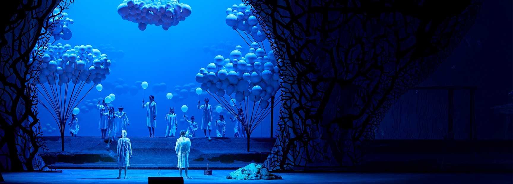 Auf der Bühne in blauer Beleuchtung stehen mehrere Personen in weißen Gewändern, die Ballons halten. Im Hintergrund sind große, wolkenähnliche Strukturen aus Ballons zu sehen. Eine Figur in einem weißen Mantel ist vorne mit dem Rücken zur Bühne gewandt.
