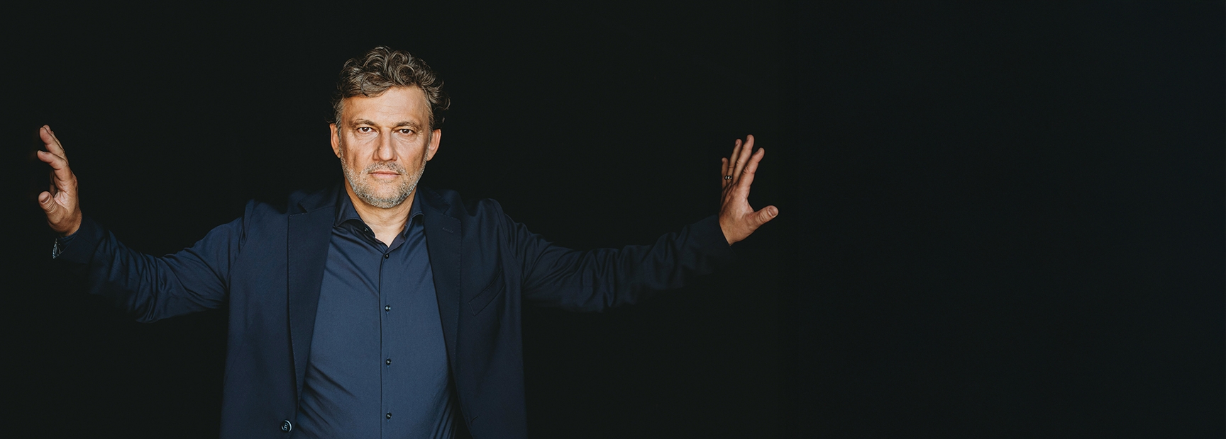 Ein Mann mit kurzen, lockigen Haaren und Bart steht vor einem dunklen Hintergrund. Er trägt ein dunkles Sakko und ein Hemd. Seine Arme sind weit ausgestreckt, als ob er eine Ausdrucksbewegung macht. Sein Gesicht zeigt einen ernsten Ausdruck.