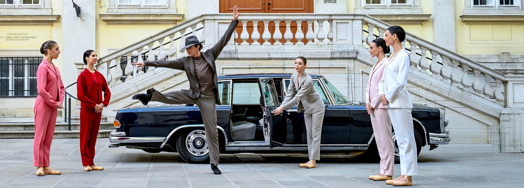 Eine Gruppe von fünf Tänzern in stilvollen Anzügen steht vor einer eleganten Limousine. Ein Tänzer führt einen dynamischen Sprung aus, während eine Tänzerin die Autotür öffnet. Die Szene spielt in einem urbanen Umfeld mit historischer Architektur im Hintergrund.