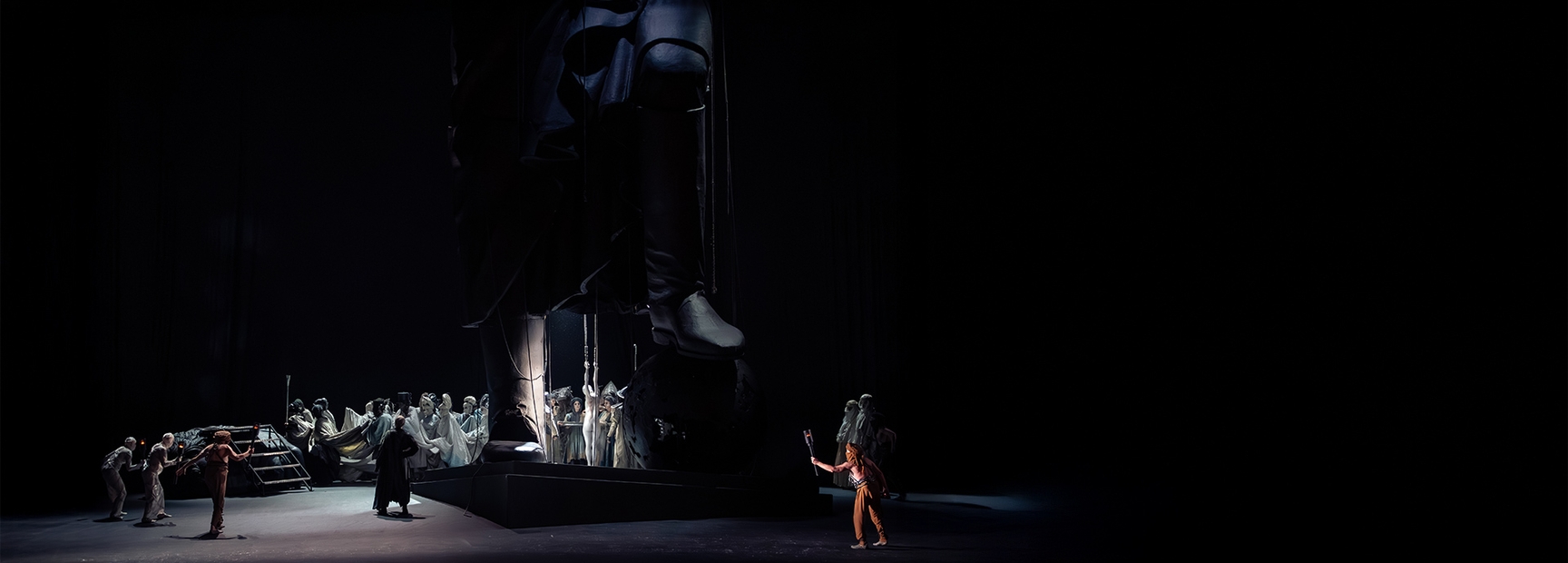 Eine dramatische Bühnenaufführung zeigt eine massive Statue im Hintergrund, während mehrere Figuren in Kostümen im Vordergrund agieren. Eine Figur mit einem Werkzeug in der Hand wirkt aktiv, während die anderen in einer Gruppendynamik interagieren. Die Szene vermittelt eine Atmosphäre von Spannung und Konflikt.