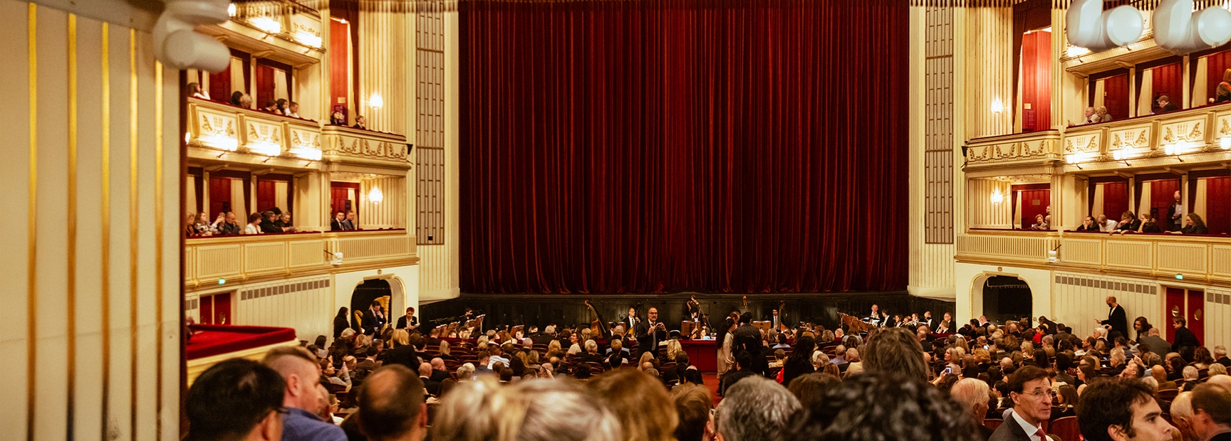 Eine klassisch gestaltete Theateraufführung ist im Gange. Der Zuschauerraum ist mit Menschen gefüllt, während der Hauptbühnenvorhang aus rotem Samt vor der leeren Bühne schließt. An den Seiten sind Kassen und logenartige Sitze sichtbar. Die Atmosphäre ist gespannt und erwartungsvoll.