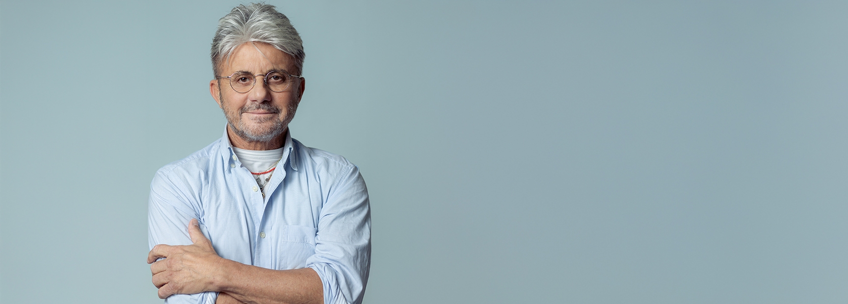 Ein Mann mittleren Alters steht lächelnd vor einem einfarbigen Hintergrund. Er trägt eine hellblaue Hemd und hat graue Haare. Seine Arme sind vor der Brust verschränkt, und er trägt eine Brille. Sein Ausdruck wirkt freundlich und selbstbewusst.