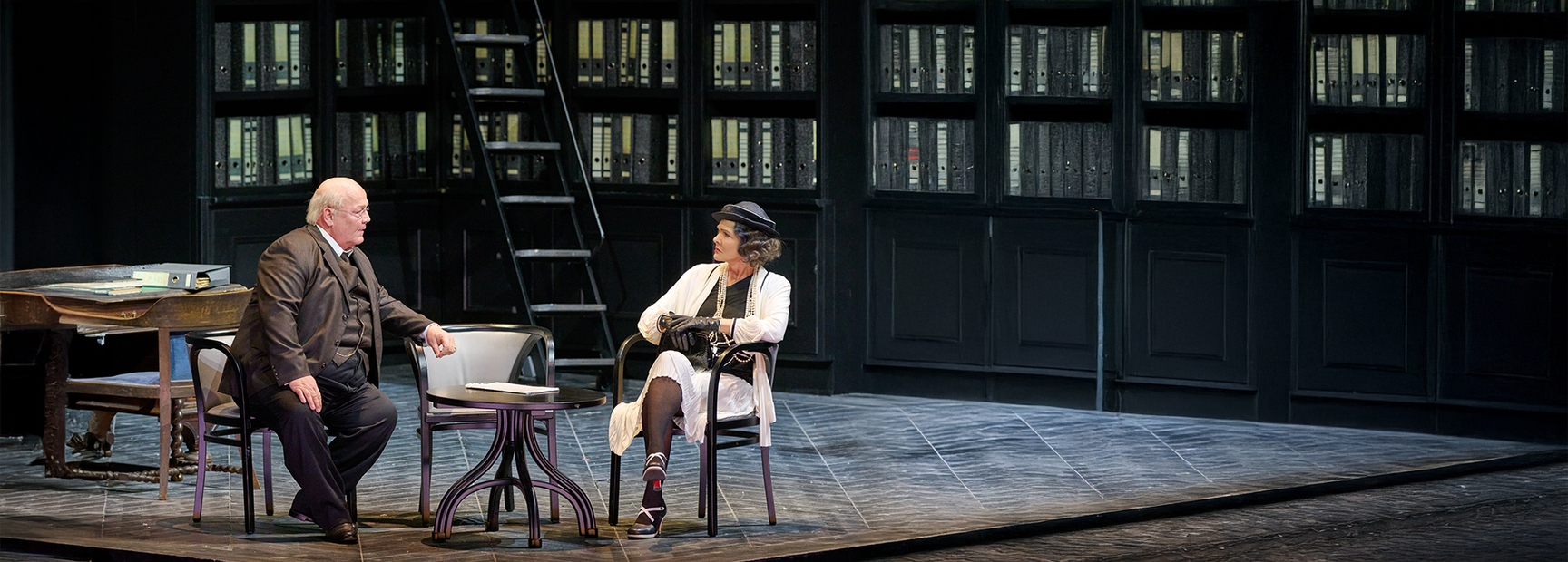 The image depicts two characters seated on a stage, engaged in conversation. One character, a man in a dark suit, gestures with his hand, while the other, a woman in a white outfit and hat, listens attentively. Behind them is a wall of shelves lined with files.