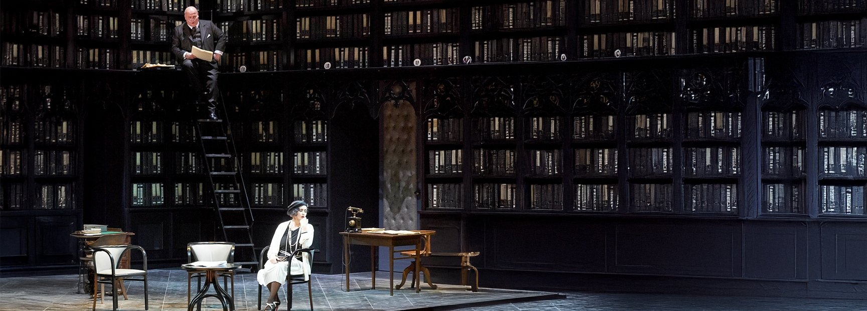 Die Szene zeigt eine Bibliothek mit hohen Regalen voller Bücher. Oben auf einer Leiter steht eine Person mit Unterlagen, während eine weitere Person an einem Tisch sitzt, nachdenklich auf ihren Handrücken gestützt. Ihr Blick ist in die Ferne gerichtet.