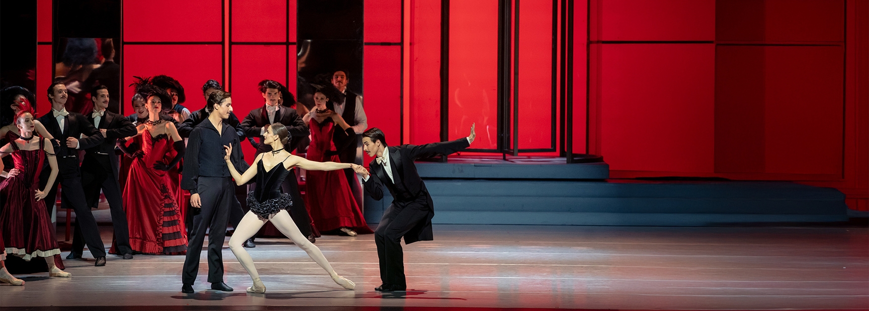 A group of dancers performs on stage, featuring a male dancer in a black suit and a female dancer in a black dress, engaged in an expressive duet. The backdrop is a striking red, enhancing the dramatic atmosphere of the performance. Other dancers observe in elegant costumes.