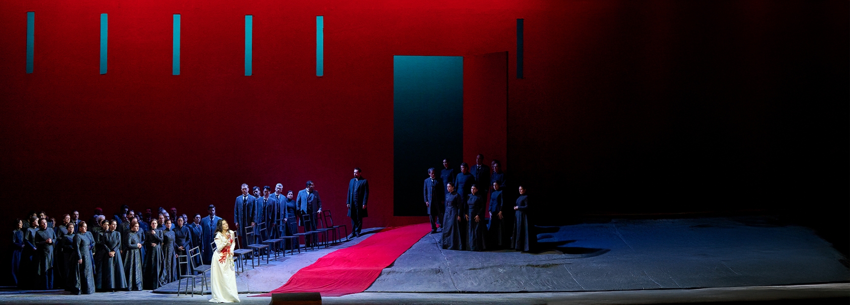 Eine Sängerin in einem weißen Kleid mit roten Details steht im Vordergrund einer Bühne. Hinter ihr versammeln sich viele Darsteller in dunkler Kleidung. Die Szenerie ist von intensivem Rot dominiert, mit einer großen, dunklen Tür im Hintergrund, was eine dramatische Atmosphäre schafft.