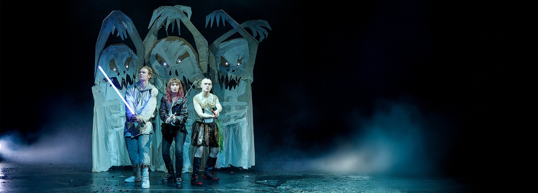 Drei Charaktere auf einer Bühne stehen zusammen, während sie nach vorne schauen. Einer hält ein Lichtschwert, während ein anderer ein Schwert hält. Im Hintergrund sind schattenhafte Gestalten zu sehen, die eine bedrohliche Atmosphäre schaffen.