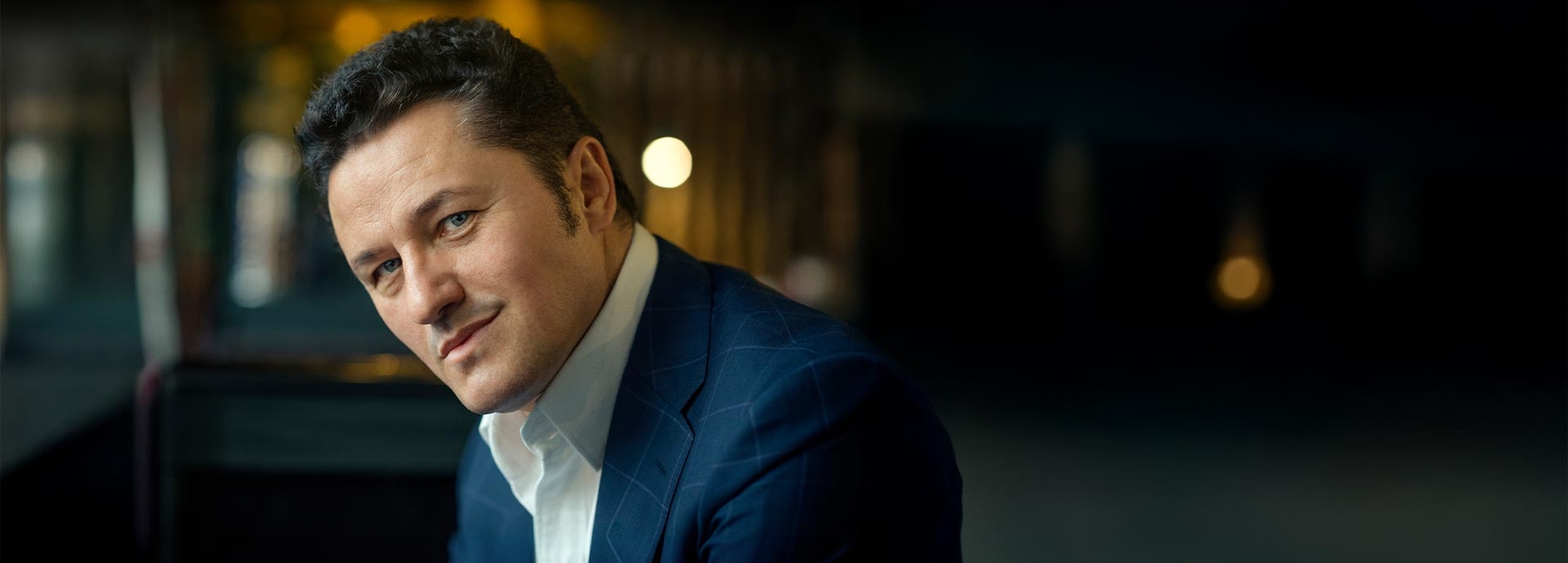 Ein Mann in einem blauen Anzug schaut direkt in die Kamera. Er hat dunkles, lockiges Haar und einen leicht lächelnden Gesichtsausdruck. Der Hintergrund ist unscharf, mit sanften Lichtern, die einen eleganten Eindruck vermitteln.