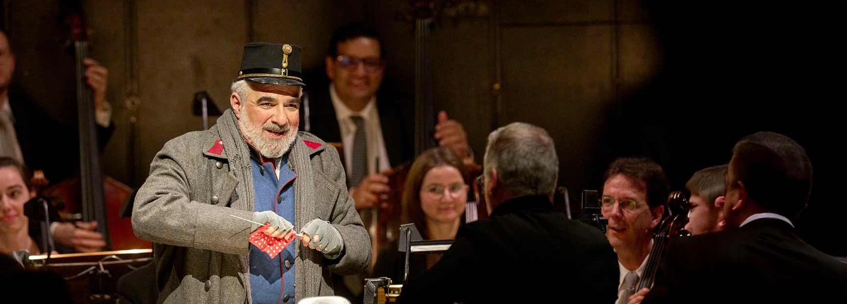 Ein älterer Mann in einem grauen Mantel und einer Schirmmütze steht auf einer Bühne und hält ein rotes Tuch. Er lächelt, während er von einem Orchester umgeben ist, das gerade spielt. Die Szene vermittelt eine festliche und musikalische Atmosphäre.