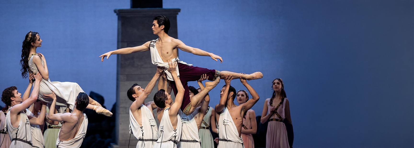 A ballet performance showcases dancers in elegant costumes. A male dancer holds a female dancer above his head, while several other male dancers support him, creating a dynamic formation. The scene emphasizes grace and strength, set against a softly lit backdrop.