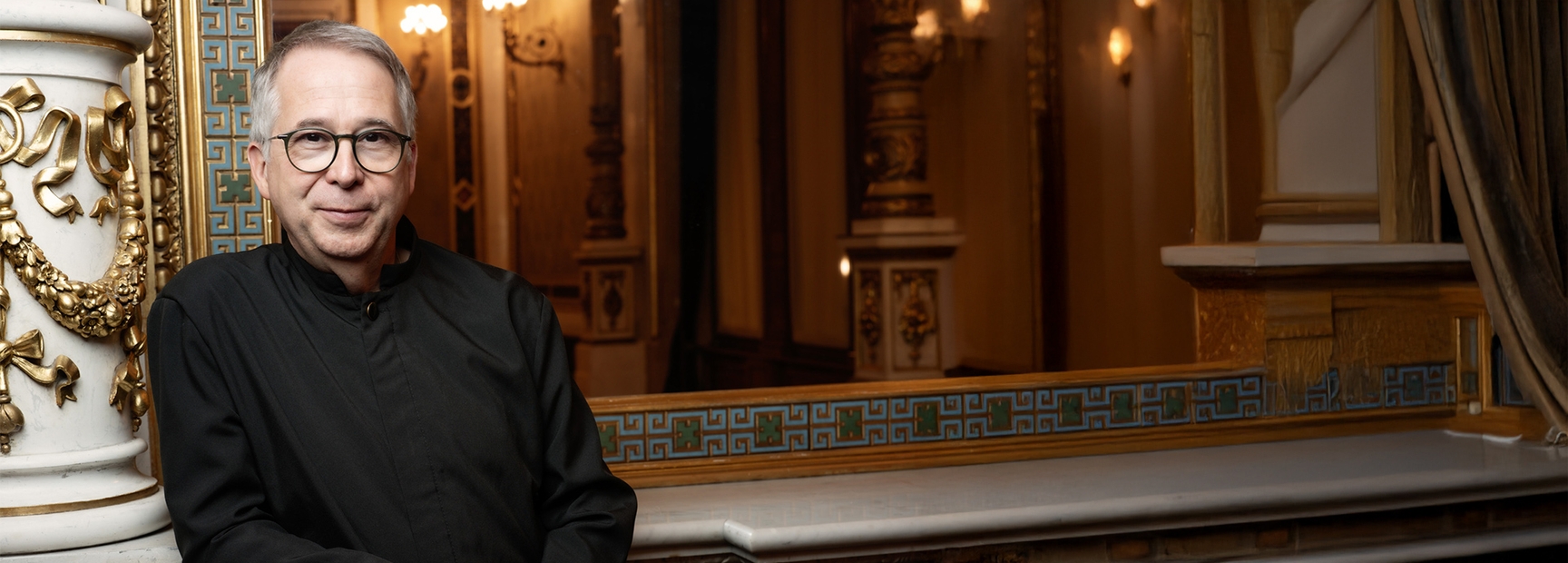 A middle-aged man with short gray hair and glasses is standing beside an ornate column, wearing a black garment. He appears confident and approachable, with a slight smile, in a sophisticated interior adorned with decorative designs and soft lighting.