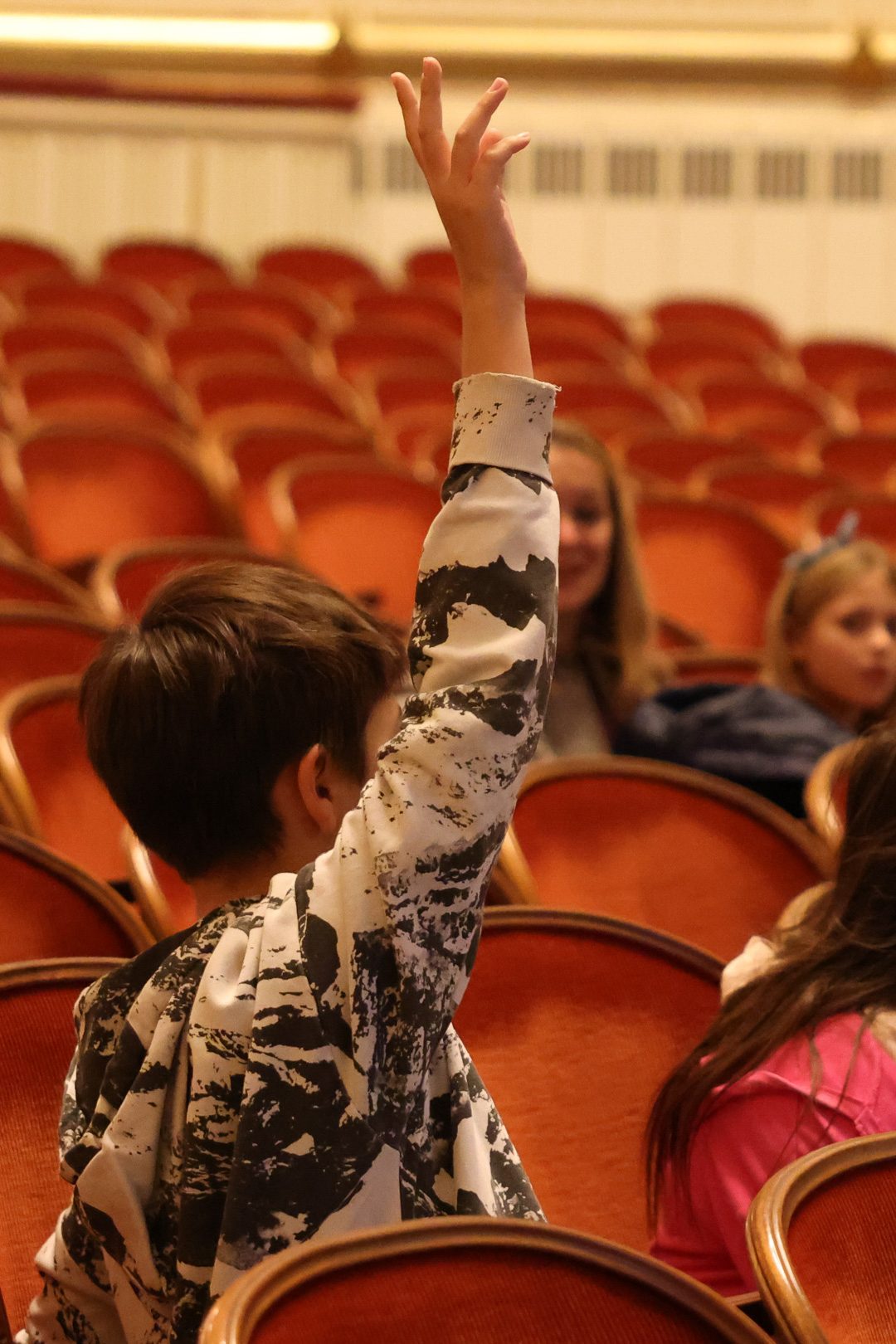 Ein Kind mit braunen Haaren hebt in einem Theatersaal die Hand. Im Hintergrund sind andere Kinder und leere rote Sitze zu sehen. Die Szene vermittelt eine Atmosphäre von Neugier und Beteiligung.