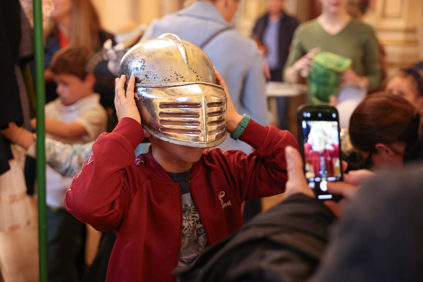 Ein Junge trägt einen glänzenden Ritterhelm und hält ihn mit beiden Händen auf seinem Kopf. Im Hintergrund sind Menschen und eine belebte Umgebung zu sehen. Es wirkt, als würde der Junge Spaß haben und wird möglicherweise fotografiert.