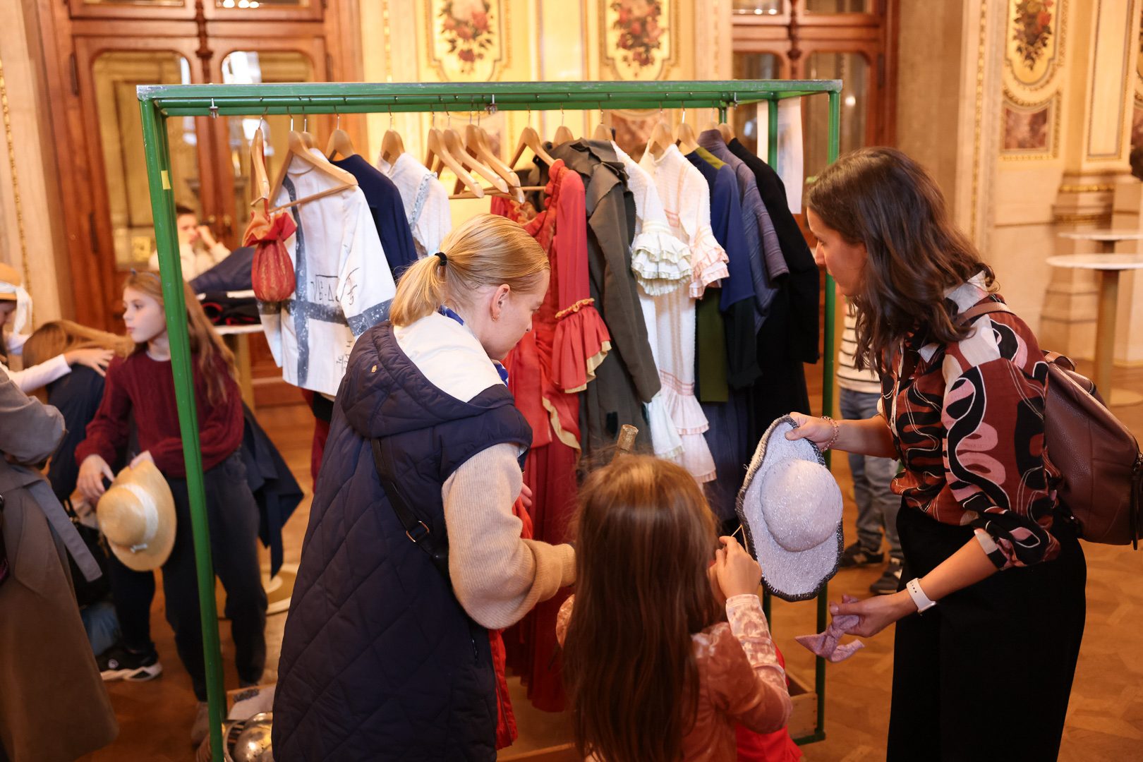 Zwei Frauen und ein Mädchen betrachten Kleidung an einem Kleiderständer. Der Ständer ist mit verschiedenen Kleidungsstücken in bunten Farben bestückt. Eine Frau hält einen Hut, während das Mädchen an ihrer Seite interessiert schaut. Der Hintergrund zeigt eine dekorative Innenarchitektur.