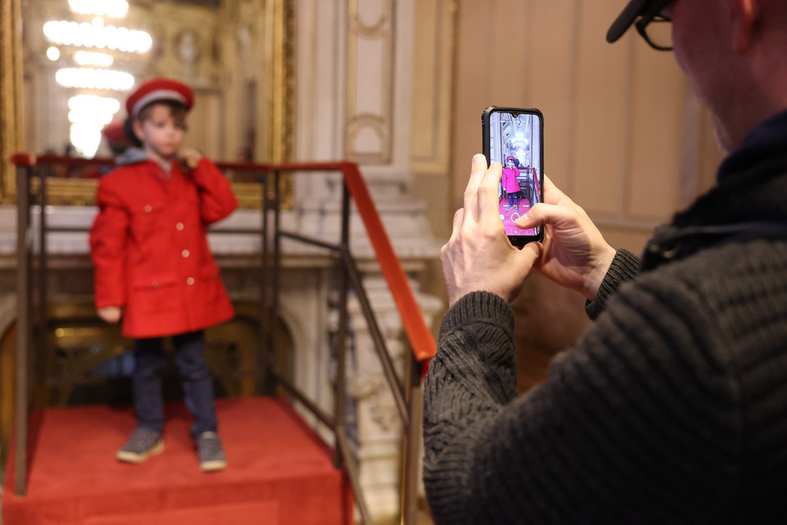 Ein Kind in einem roten Mantel und einer Mütze steht auf einer kleinen Plattform und posiert. Eine Person im Vordergrund hält ein Smartphone, um ein Foto von dem Kind zu machen. Der Hintergrund zeigt eine prächtige Innenarchitektur.