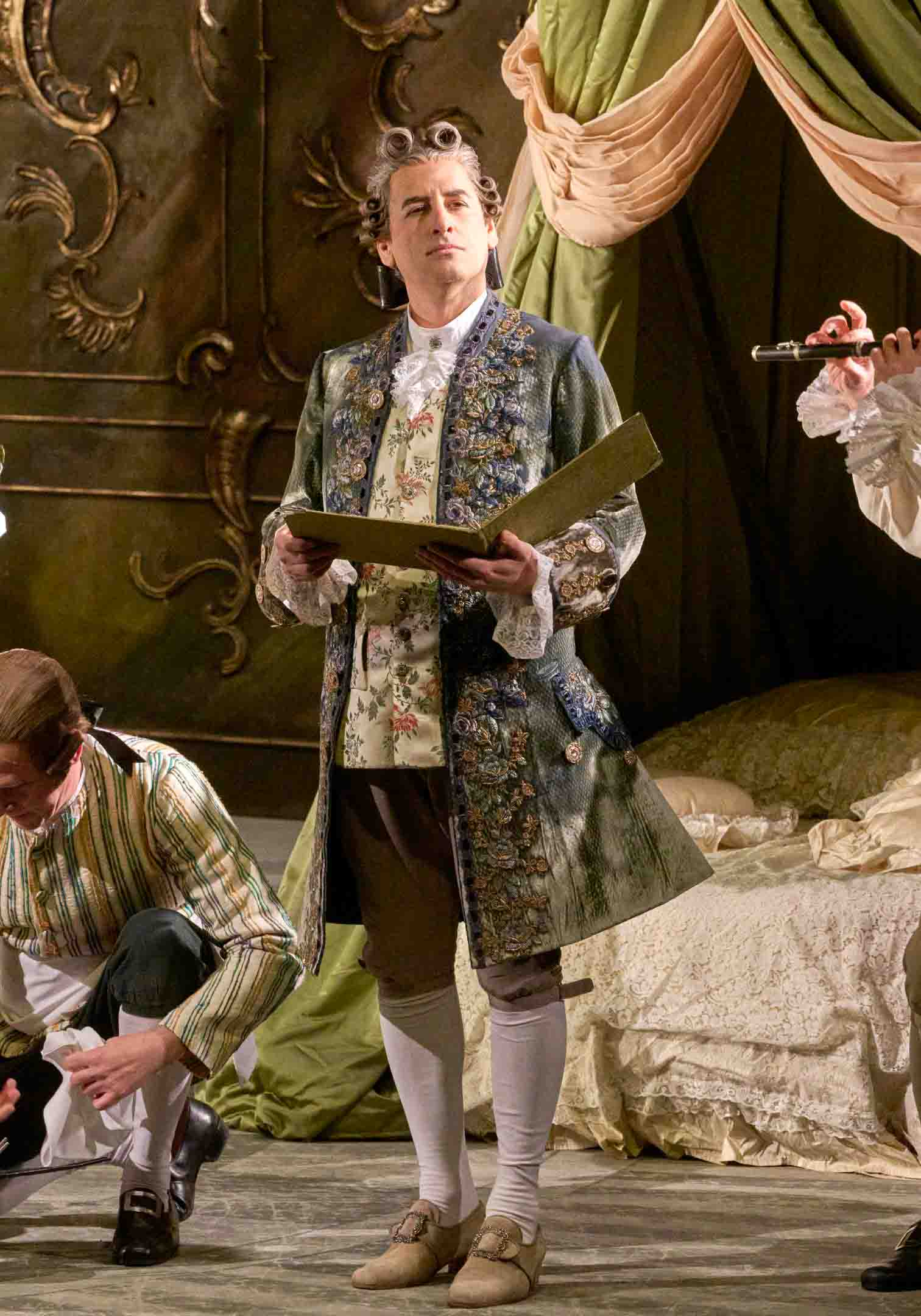 Ein Mann in historischer Kleidung mit lockigem Haar steht im Zentrum der Szene und hält ein geöffnetes Buch. Im Hintergrund sind weitere Personen und aufwendige Stoffe sichtbar, die eine theaterähnliche Kulisse schaffen. Der Mann scheint zu lesen oder Anweisungen zu geben.