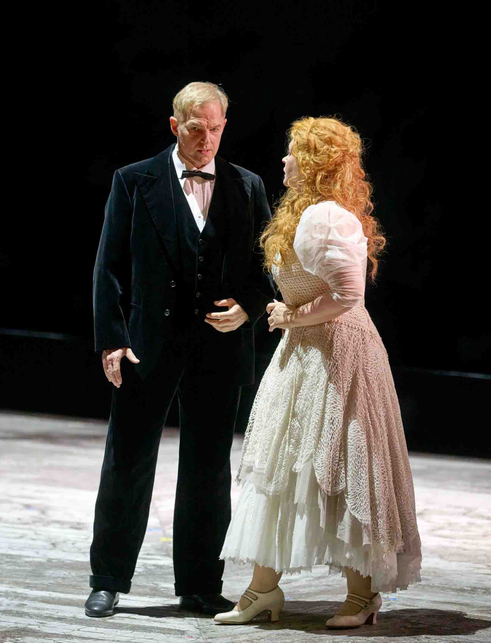 Ein Mann in einem schwarzen Anzug steht neben einer Frau in einem eleganten, weißen Kleid. Sie haben einen intensiven Blickkontakt und wirken in einem ernsten Gespräch vertieft. Der Hintergrund ist dunkel, was die beiden Protagonisten hervorhebt.