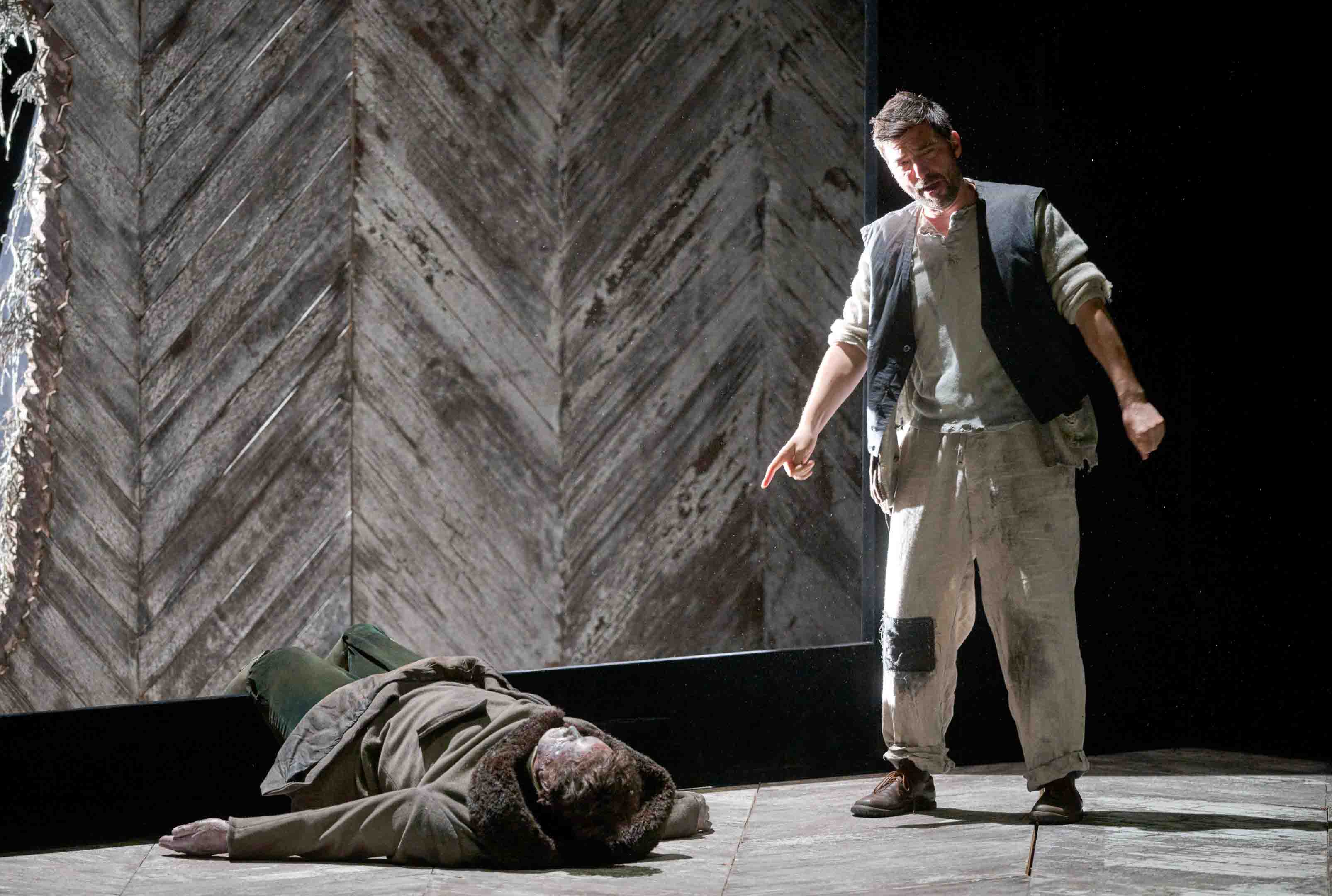 Ein stehender Mann mit zerrissener Kleidung zeigt auf einen am Boden liegenden, in einem Mantel gehüllten Körper. Die Szene spielt in einem düsteren, minimalistischen Setting mit einem hölzernen Hintergrund. Die Stimmung ist ernst und dramatisch.