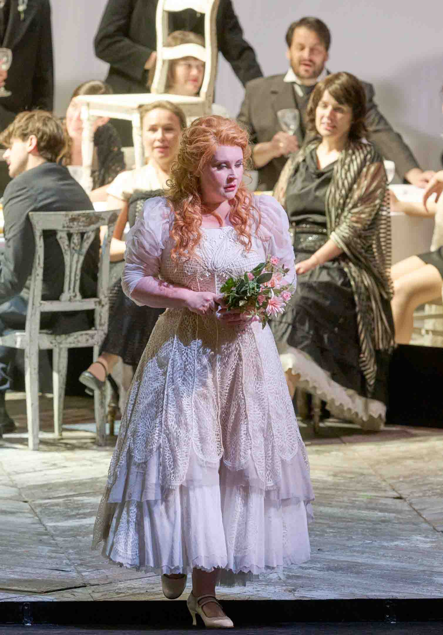 Eine Frau in einem eleganten, weißen Kleid mit Rüschen steht auf einer Bühne und hält einen Strauß Blumen. Im Hintergrund sind weitere Personen in formeller Kleidung an einem Tisch versammelt, die eine Feier oder ein gesellschaftliches Ereignis zu genießen scheinen.