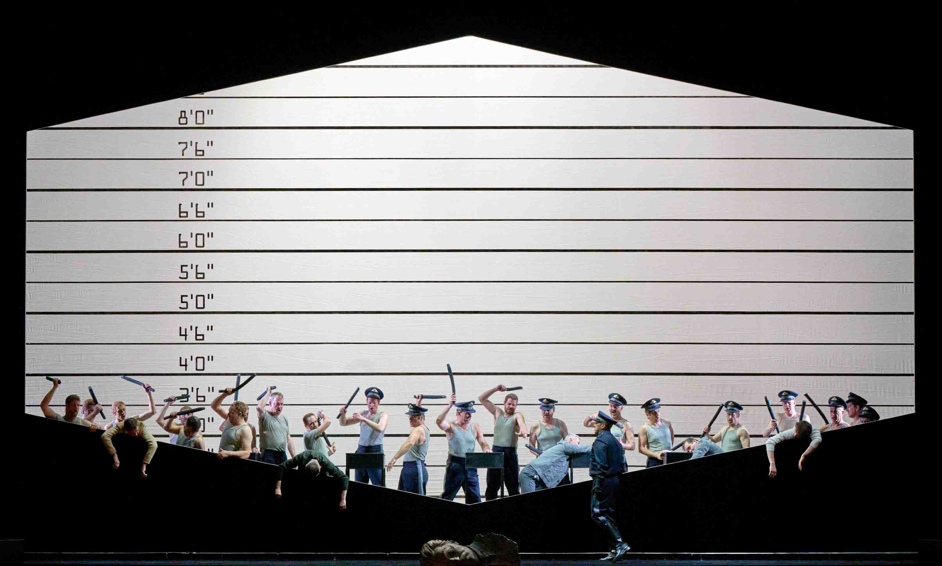 Eine Gruppe von darstellenden Personen steht auf einer Bühne, umgeben von einer großen, grafischen Wand mit Höhenmarkierungen. Einige Personen halten Stöcke oder Schlagwerkzeuge. Die Szene vermittelt eine dynamische, möglicherweise konfliktbeladene Stimmung, während die Akteure in verschiedenen Posen interagieren.