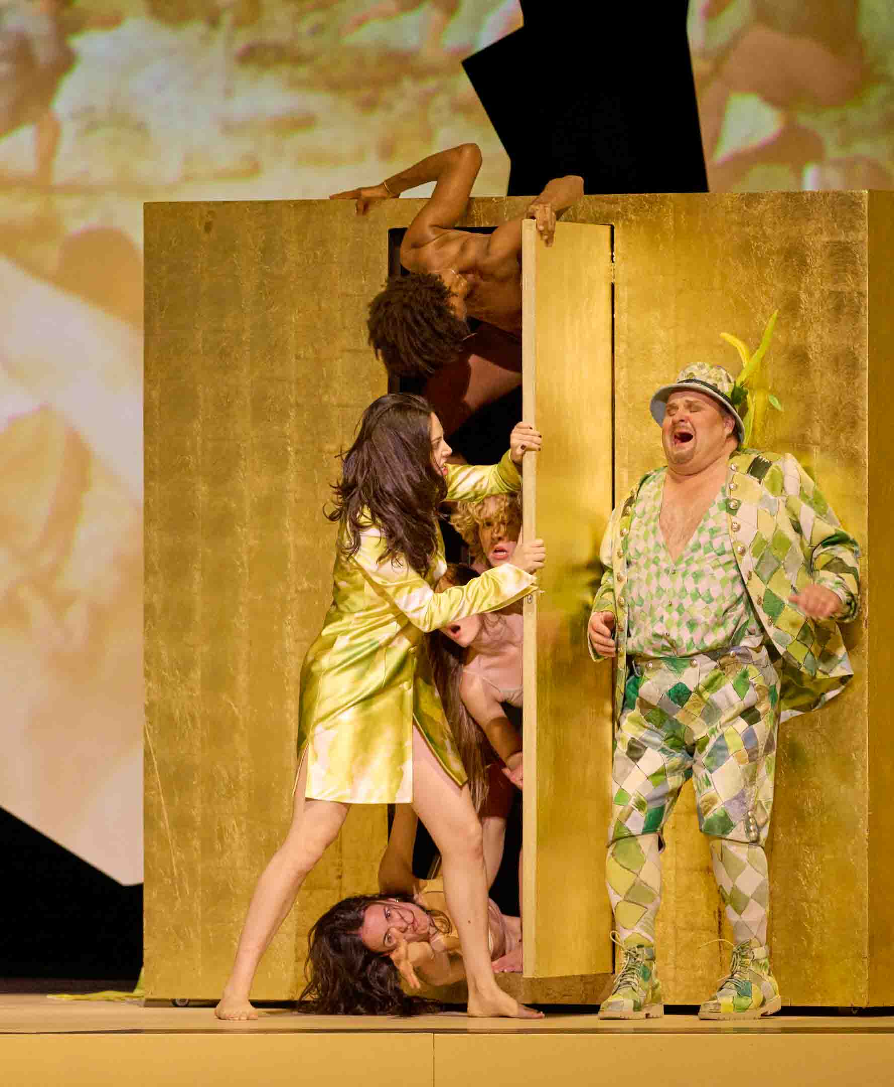 Eine Gruppe von fünf Personen agiert auf einer Bühne. Eine Frau in einem glitzernden, gelben Outfit öffnet eine goldene Tür, während eine weitere Person aus der Tür hervorspringt. Die Szene vermittelt dramatische und humorvolle Elemente einer Theateraufführung.