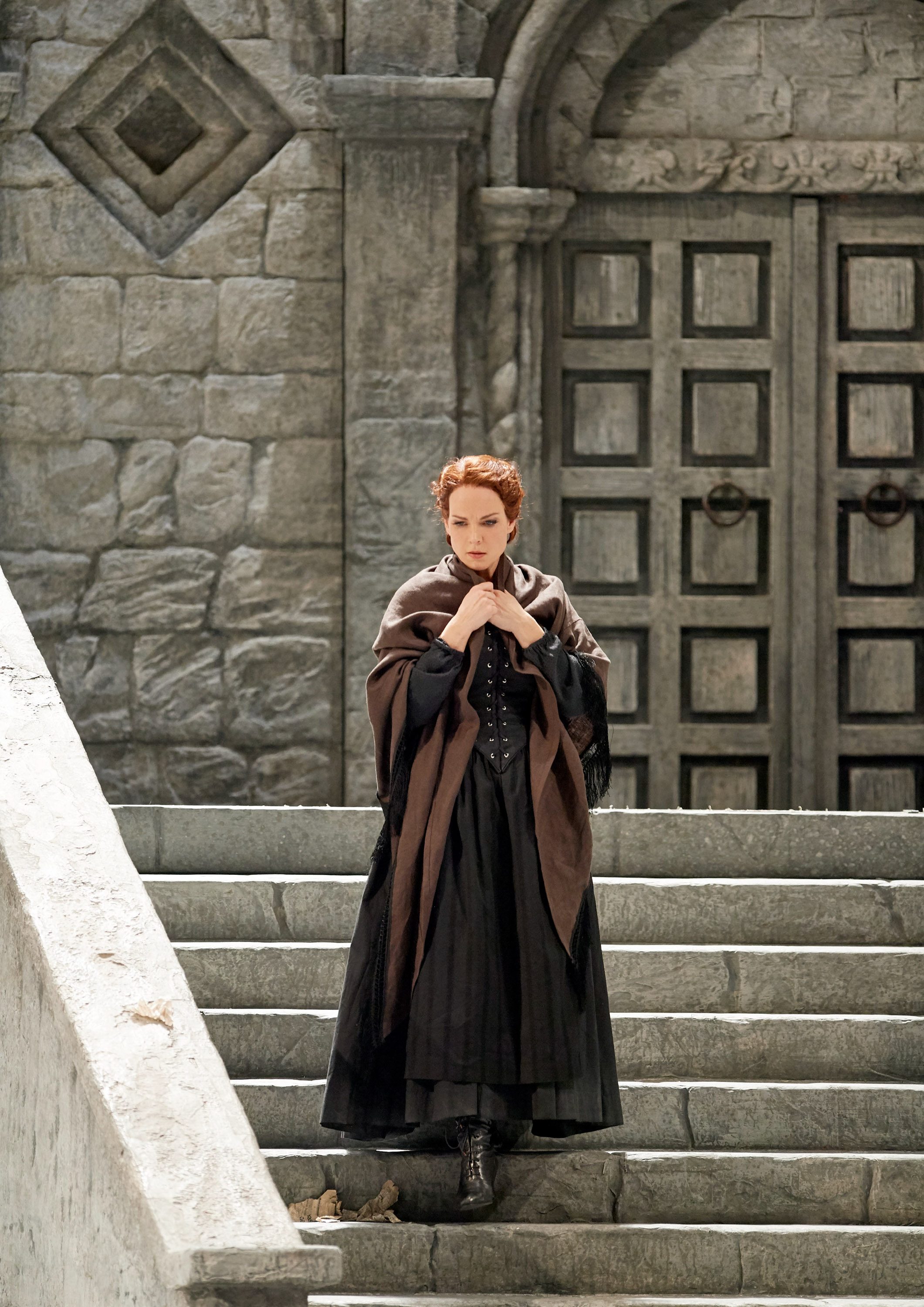 A woman stands on stone steps, wearing a long black dress and a brown shawl. She looks pensive, with her hands clasped together. Behind her is a large, intricately designed wooden door set in a grey stone wall. The scene conveys a sense of contemplation and drama.
