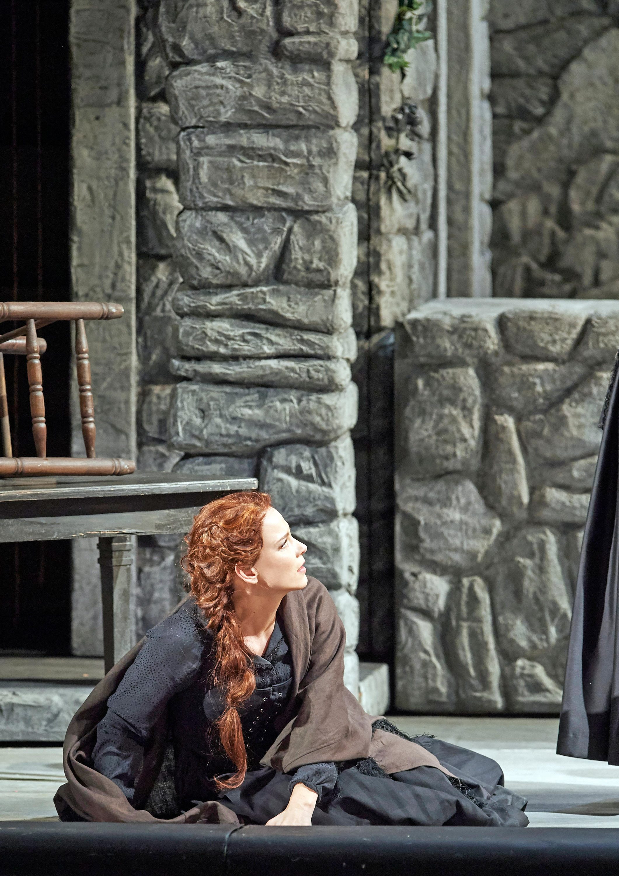 A woman with long, wavy red hair sits on the floor, dressed in a dark costume with a brown shawl. She looks upward, expressing emotion, while a stone wall and a wooden chair are visible nearby, creating a dramatic theatrical setting.