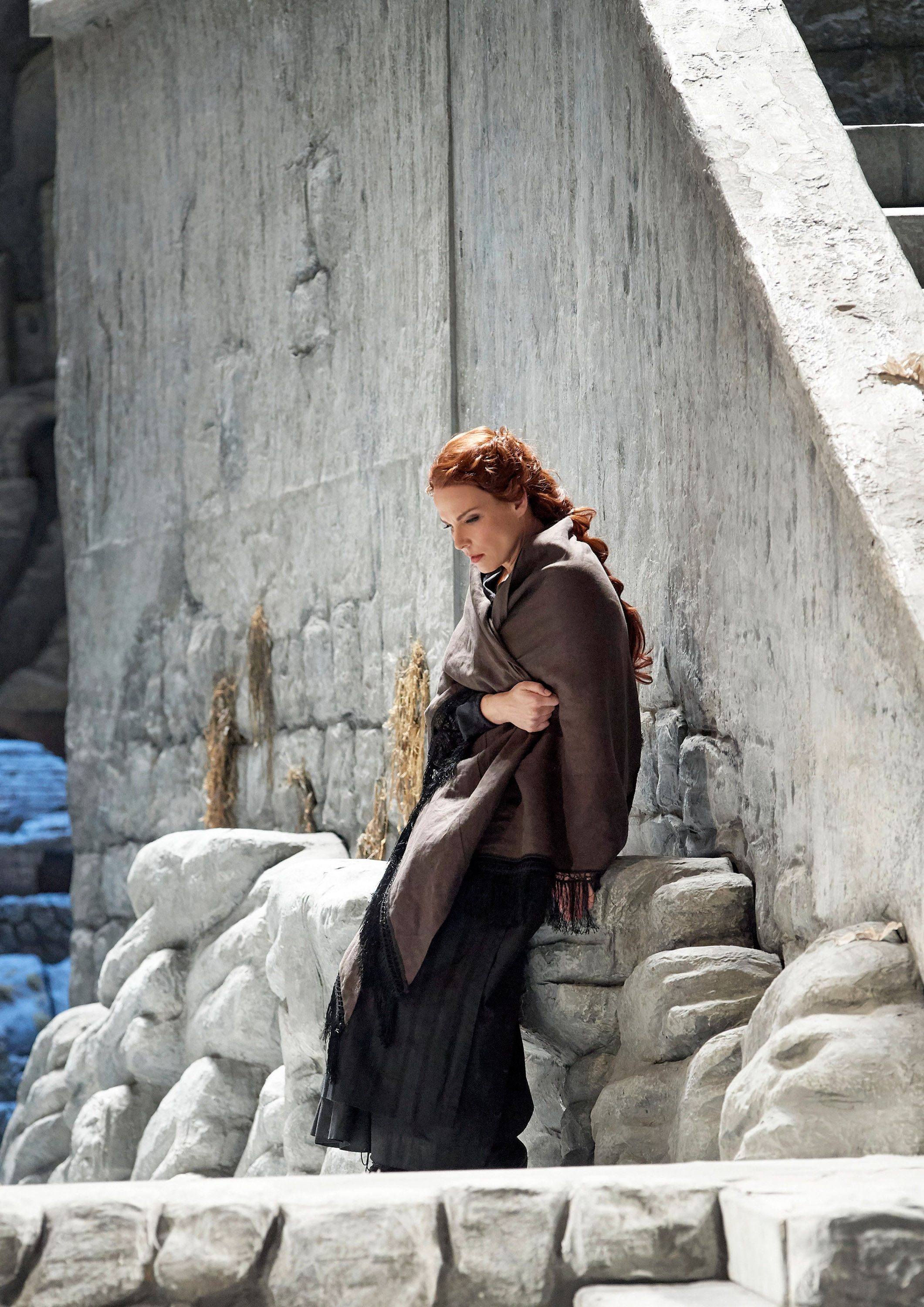 A woman with long red hair stands beside a stone wall, wearing a brown shawl and a long black skirt. She appears contemplative, looking down while hugging herself, with a somber expression on her face. The background features rough stone textures and a muted color palette.