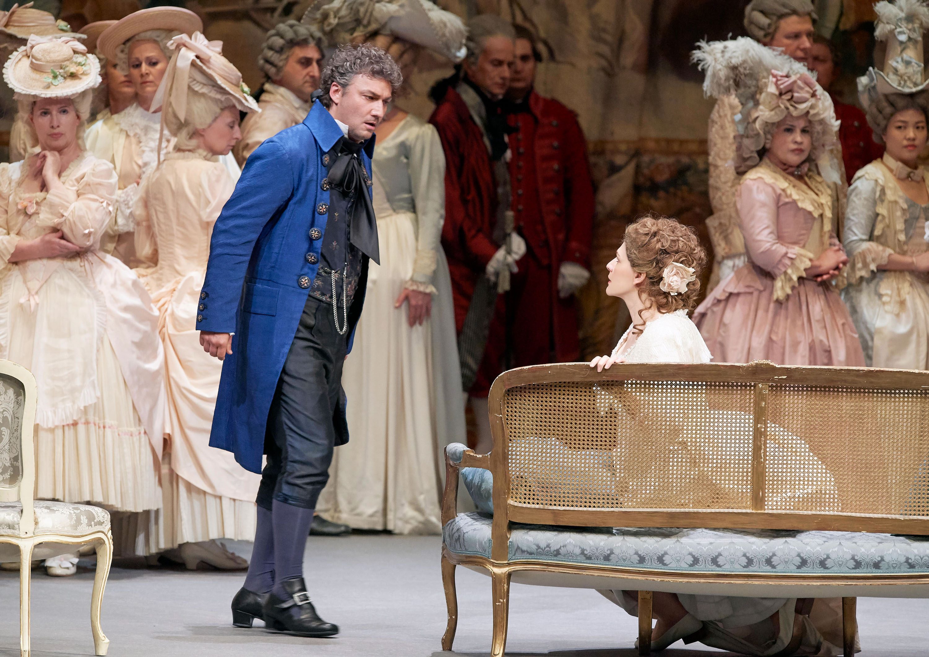 A dramatic scene from a theatrical performance, featuring a man in a blue coat confronting a woman seated on a couch. The woman, dressed in white, appears distressed. The background shows a group of elegantly dressed individuals, enhancing the emotional tension between the two main characters.