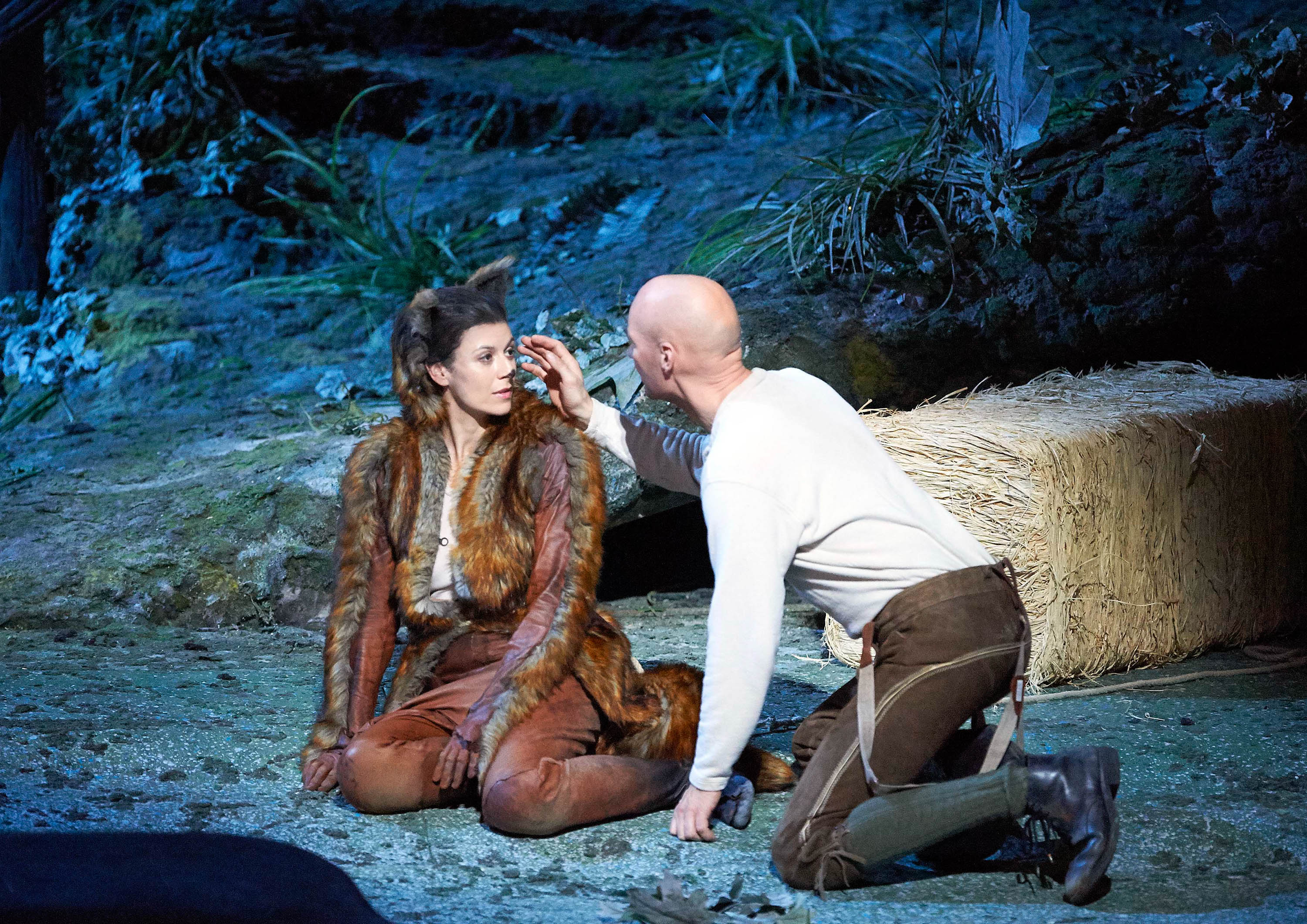A man, kneeling and dressed in a simple white shirt and brown pants, gently touches the face of a woman sitting beside him. She wears a fur costume and looks up at him with a concerned expression, set against a natural, forest-like backdrop.