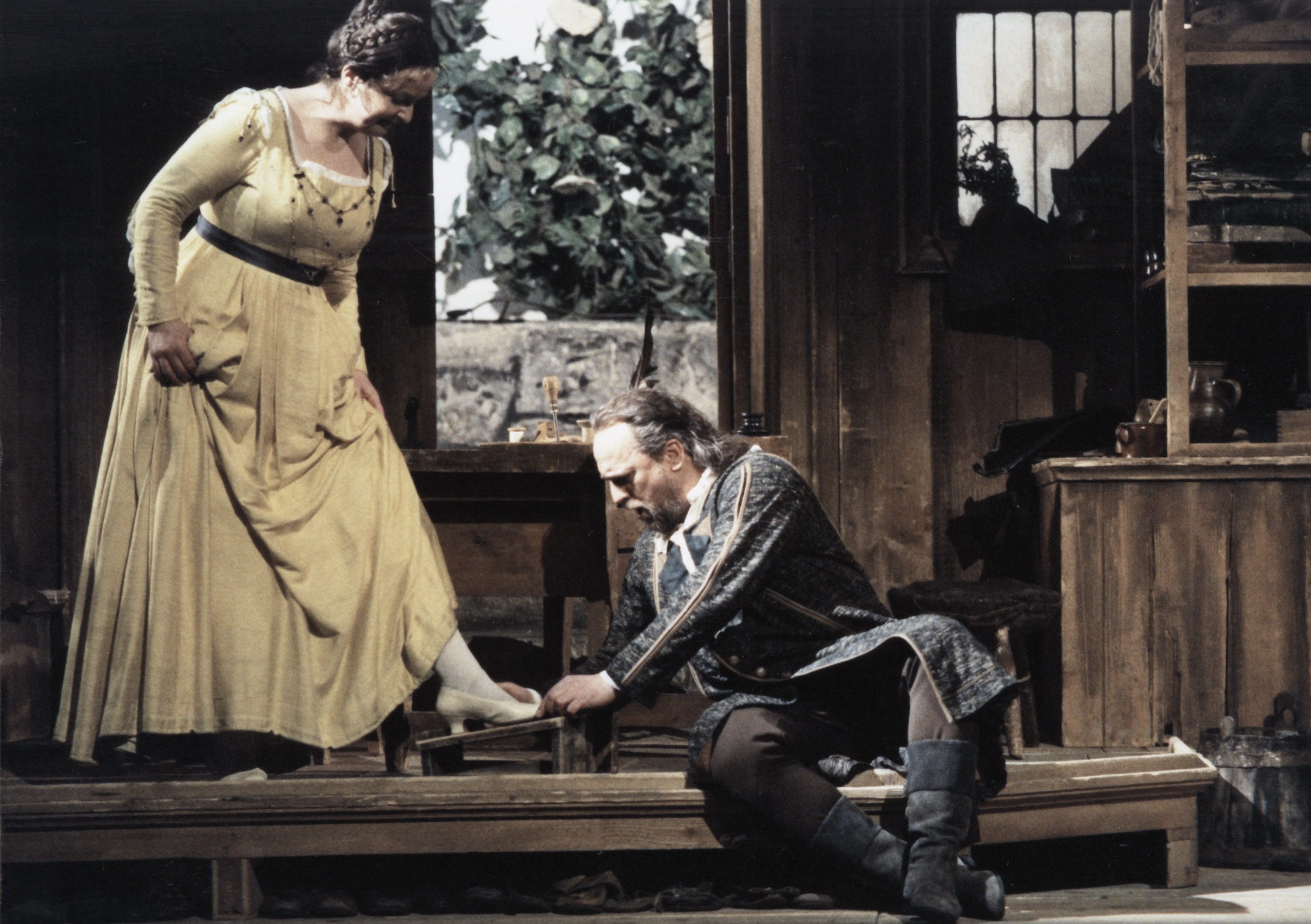 A woman in a yellow dress stands beside a seated man who is examining her foot. The setting appears to be a rustic interior, with wooden elements and a window in the background. The scene suggests a moment of care or attention.