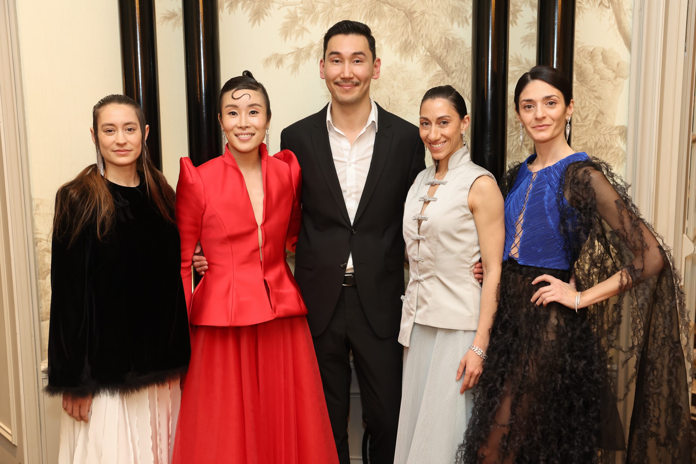 Fünf Personen stehen zusammen, die Damen tragen elegante Kleider in verschiedenen Farben und Stilen, während der Mann einen klassischen Anzug trägt. Im Hintergrund ist eine dekorative Wand mit einem floralen Muster zu sehen.