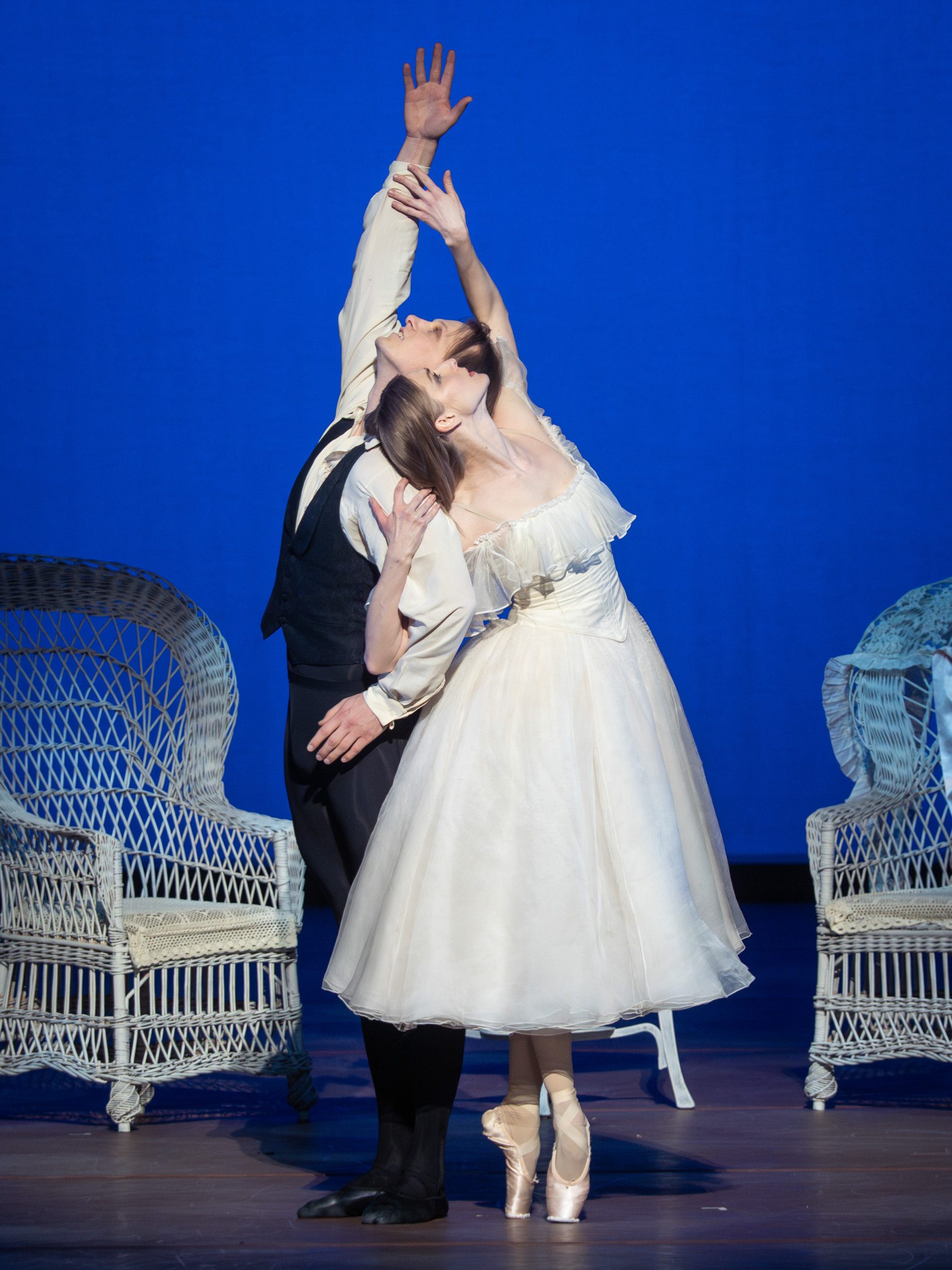 Ein männlicher Tänzer in einem schwarzen Anzug und eine Tänzerin in einem weißen Kleid führen eine elegante Pose auf der Bühne aus. Ihre Arme sind gestreckt, und sie blicken einander an, während sie in einem romantischen Moment vereint sind. Der Hintergrund ist in einem leuchtenden Blau.