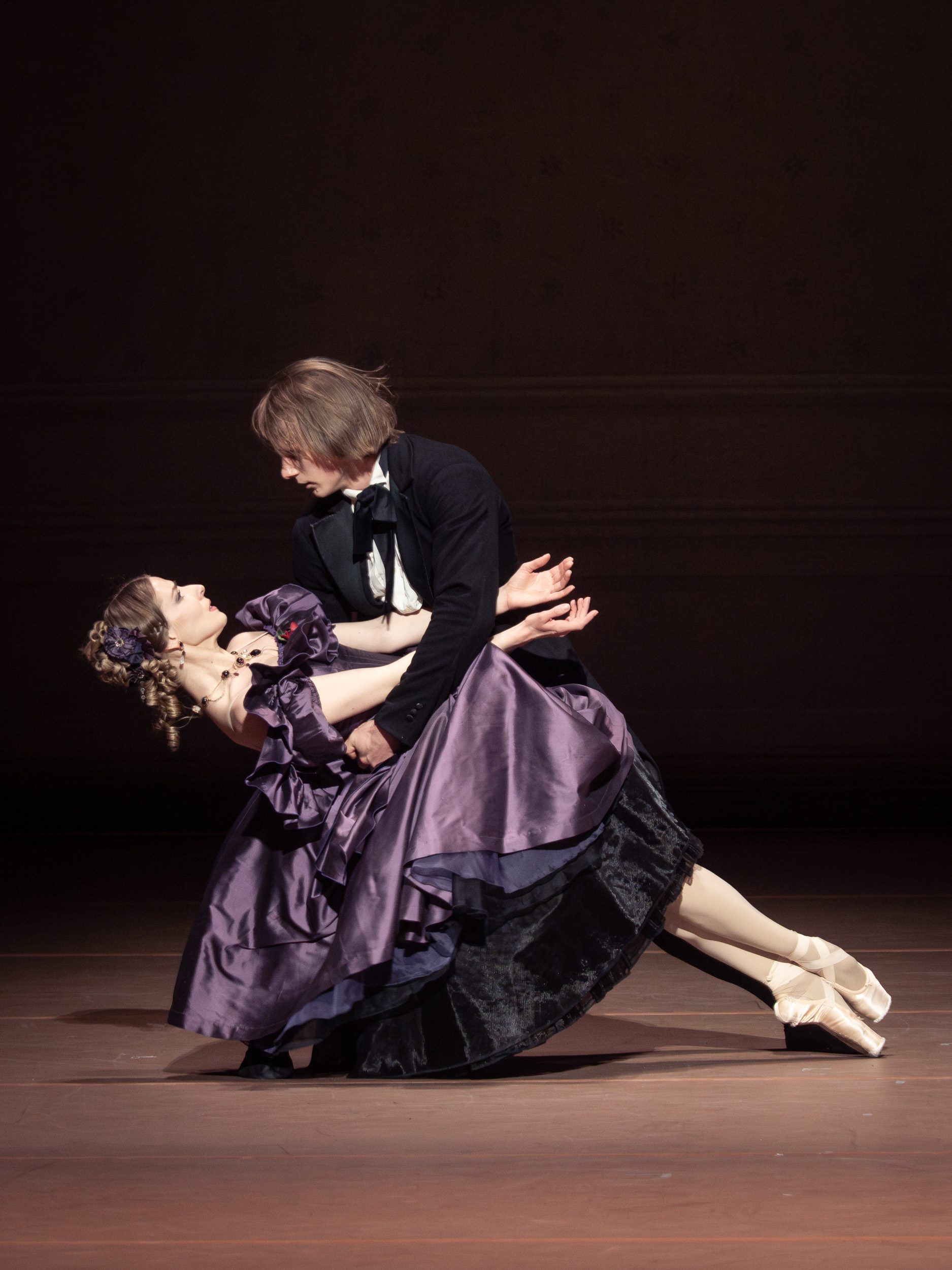 Ein männlicher und eine weibliche Tänzerin führen einen klassischen Tanz auf. Die Tänzerin trägt ein elegantes, lila Kleid und hat die Haare zu einer Frisur gesteckt. Der Tänzer hält sie in einer geduldigen, anmutigen Pose, während sie in einer schönen Darstellung miteinander verbunden sind.