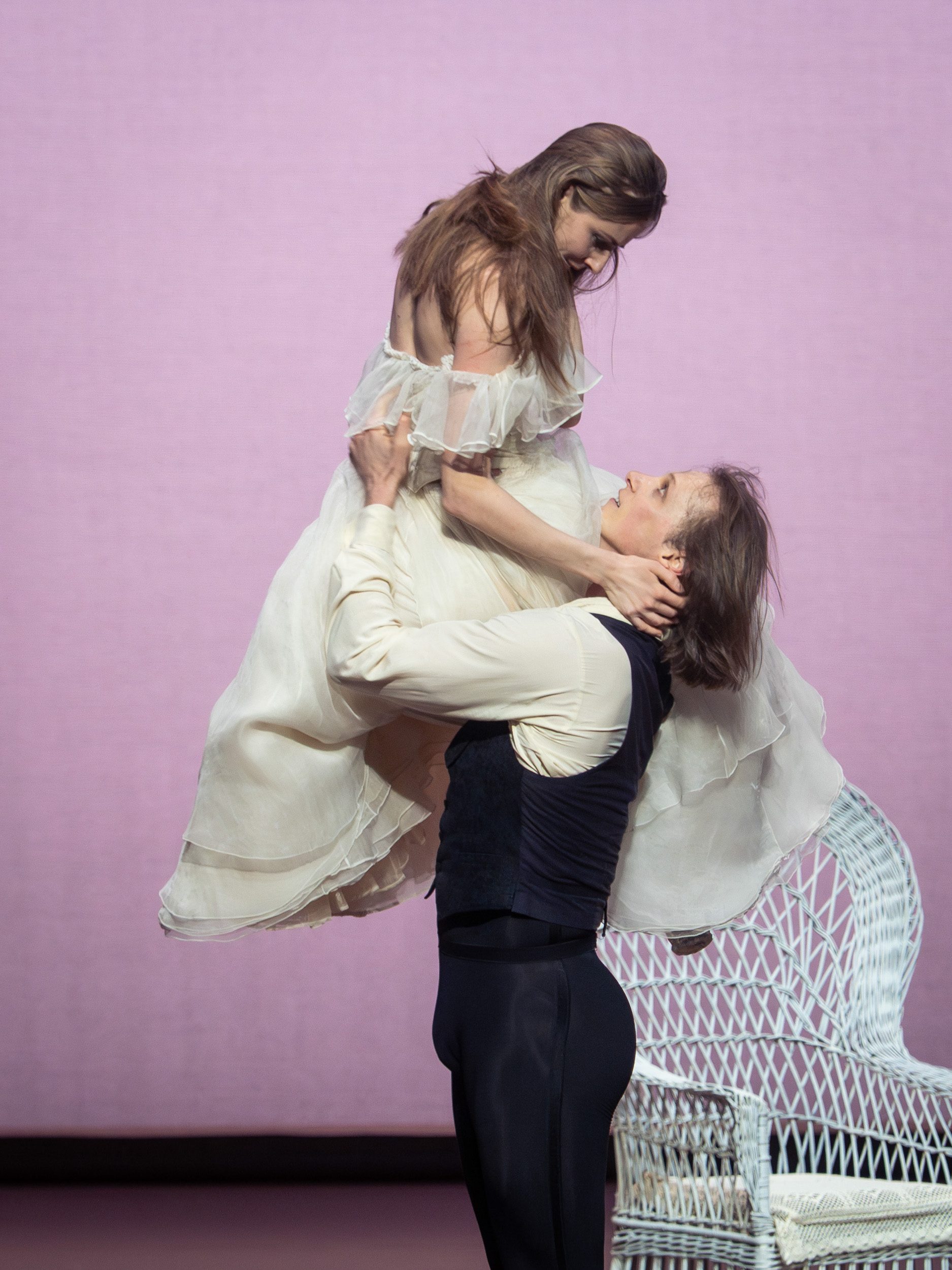 Ein männlicher Tänzer hebt eine weibliche Tänzerin, die ein elegantes, helles Kleid trägt. Die Szene strahlt Intensität und Emotion aus, während der Hintergrund in sanftem Rosa gehalten ist. Beide Tänzer zeigen eine harmonische Verbindung in ihrer Bewegung.
