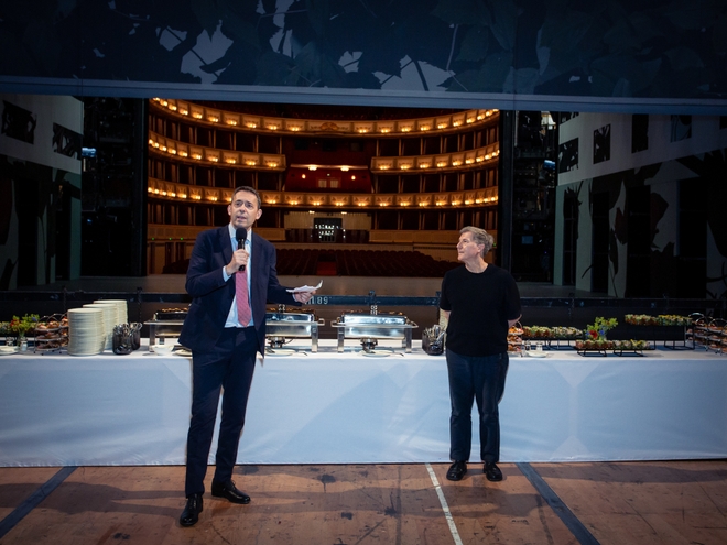Bodgan Roščić und Martin Schläpfer stehen vor einem großen Buffettisch. Im Hintergrund sieht man den Zuschauerraum der Wiener Staatsoper.