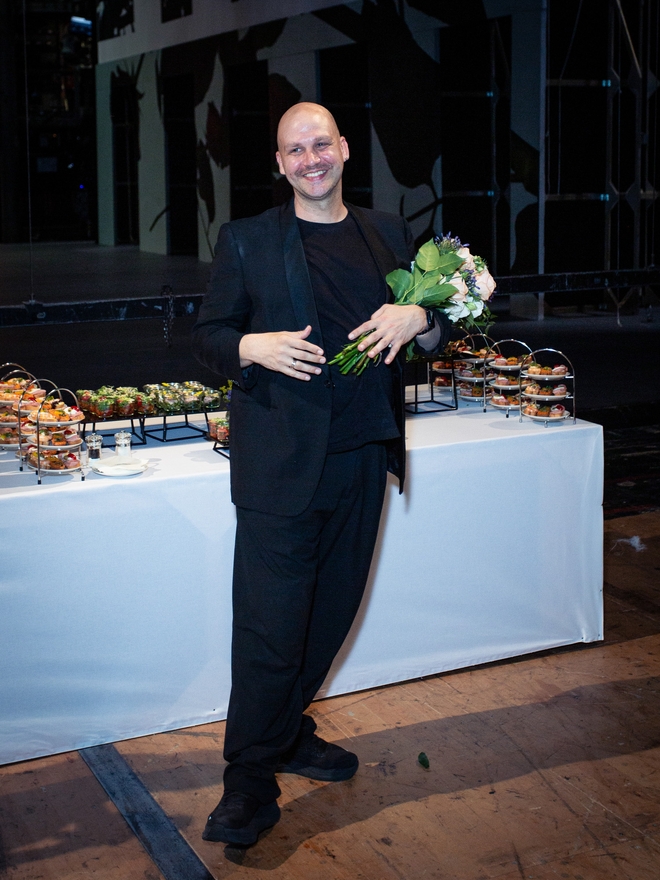 Ein Mann in schwarzem Anzug steht vor einem Buffettisch und hält einen Blumenstrauß im Arm.