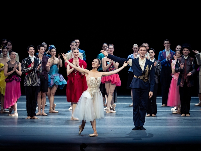 Eine Ballerina in weißem Kleid und ein Balletttänzer in einem dunklen Anzugkostüm verbeugen sich.