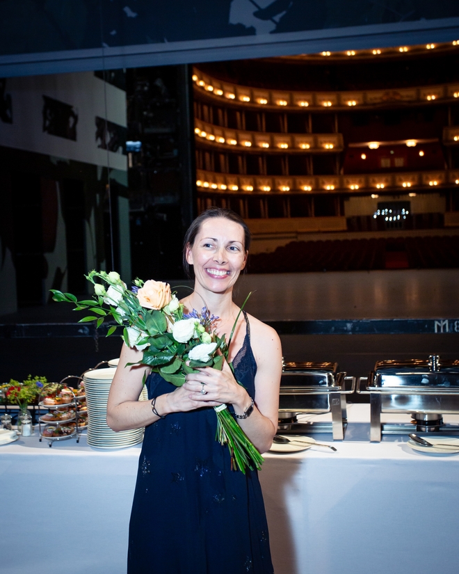Eine Frau in dunkelblauem Kleid hält einen Blumenstrauß und schaut lächelnd an der Kamera vorbei. Sie steht vor einem großen Buffettisch. Im Hintergrund ist der Zuschauerraum der Wiener Staatsoper zu sehen.