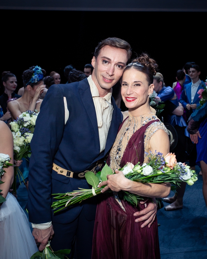 Zwei Balletttänzer*innen in dunkelblauem Anzugkostüm und einem weinroten Kleid umarmen sich halb und lächeln in die Kamera.