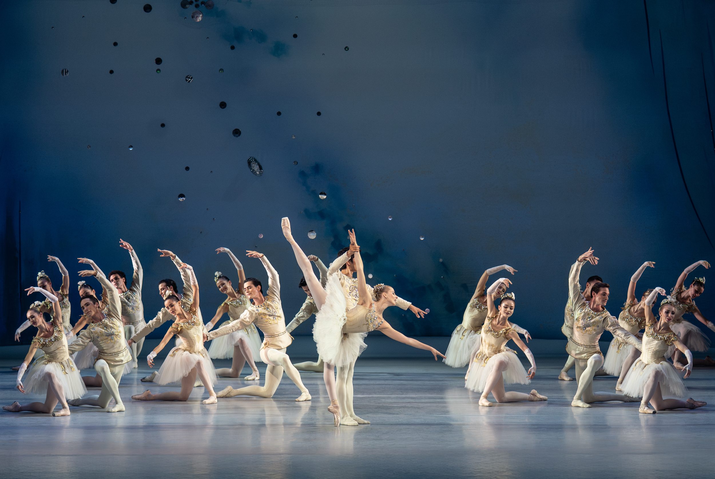 Eine Gruppe von Balletttänzern in eleganter Kleidung führt synchronisierte Bewegungen auf einer Bühne aus. Einige Tänzer heben ihre Beine und Arme, während andere in verschiedenen Posen stehen. Die Szenerie hat einen sanften, blauen Hintergrund und strahlt eine dynamische choreografische Energie aus.